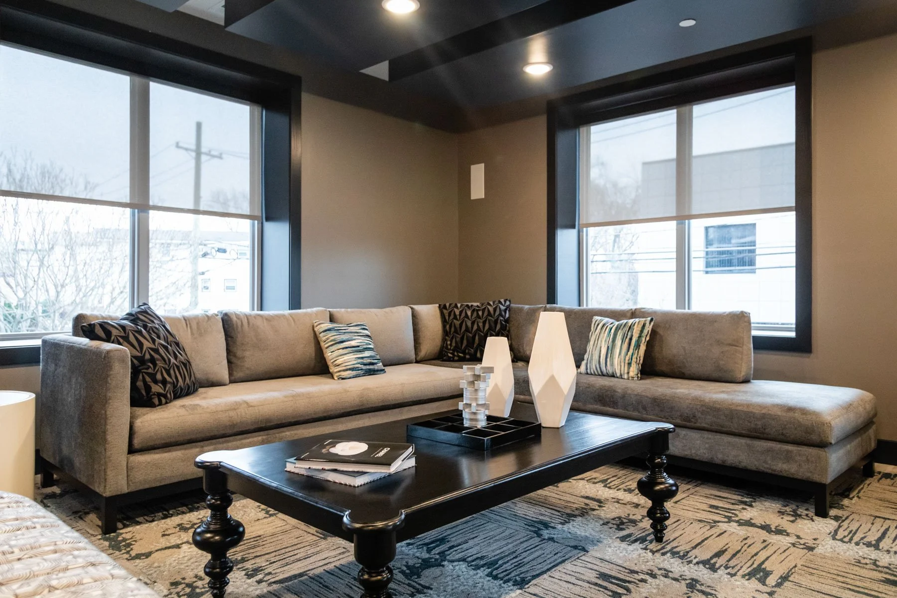 Living room with a large L-shaped beige sofa, patterned throw pillows, a dark wooden coffee table with magazines and decorative white vases, and windows with black frames showing trees and neighboring buildings outside.