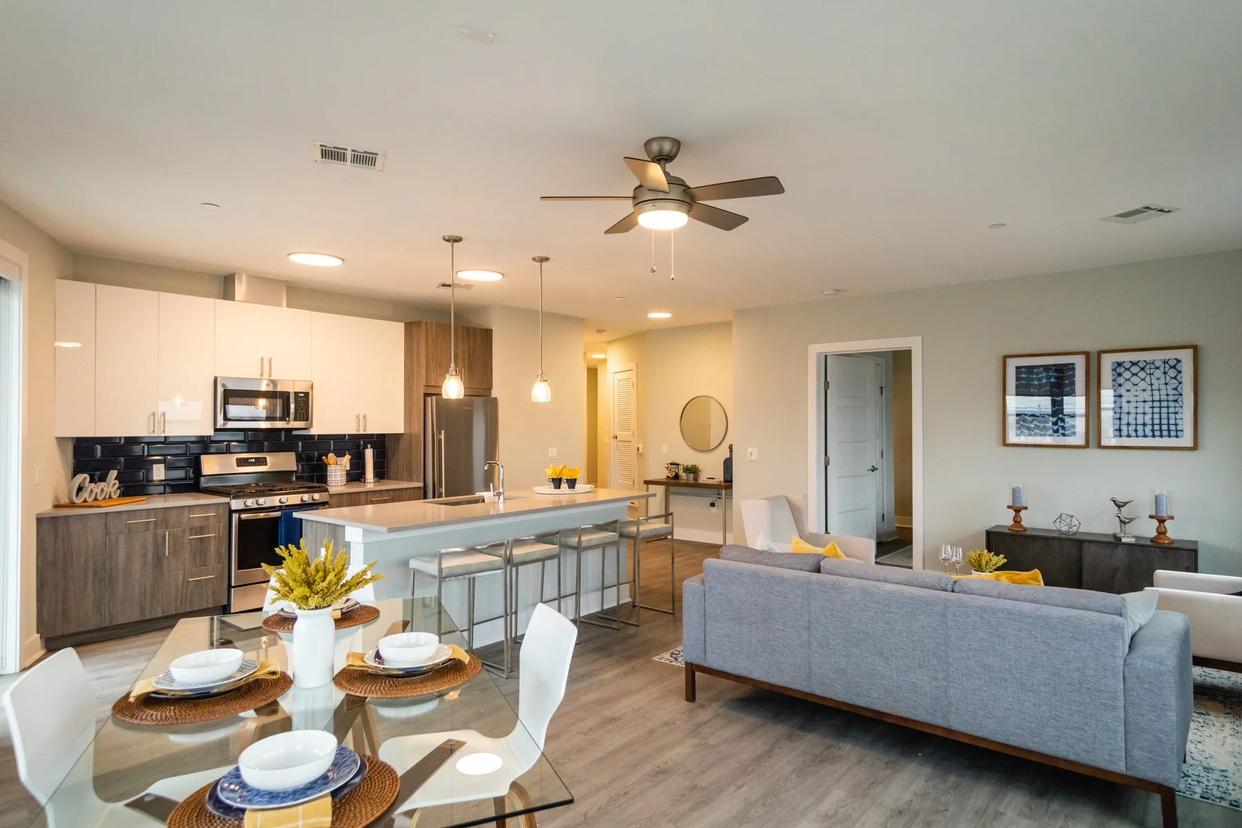 Open-concept living room and kitchen with modern decor, gray couch, dining table with four place settings, kitchen island with barstools, and stainless steel appliances.