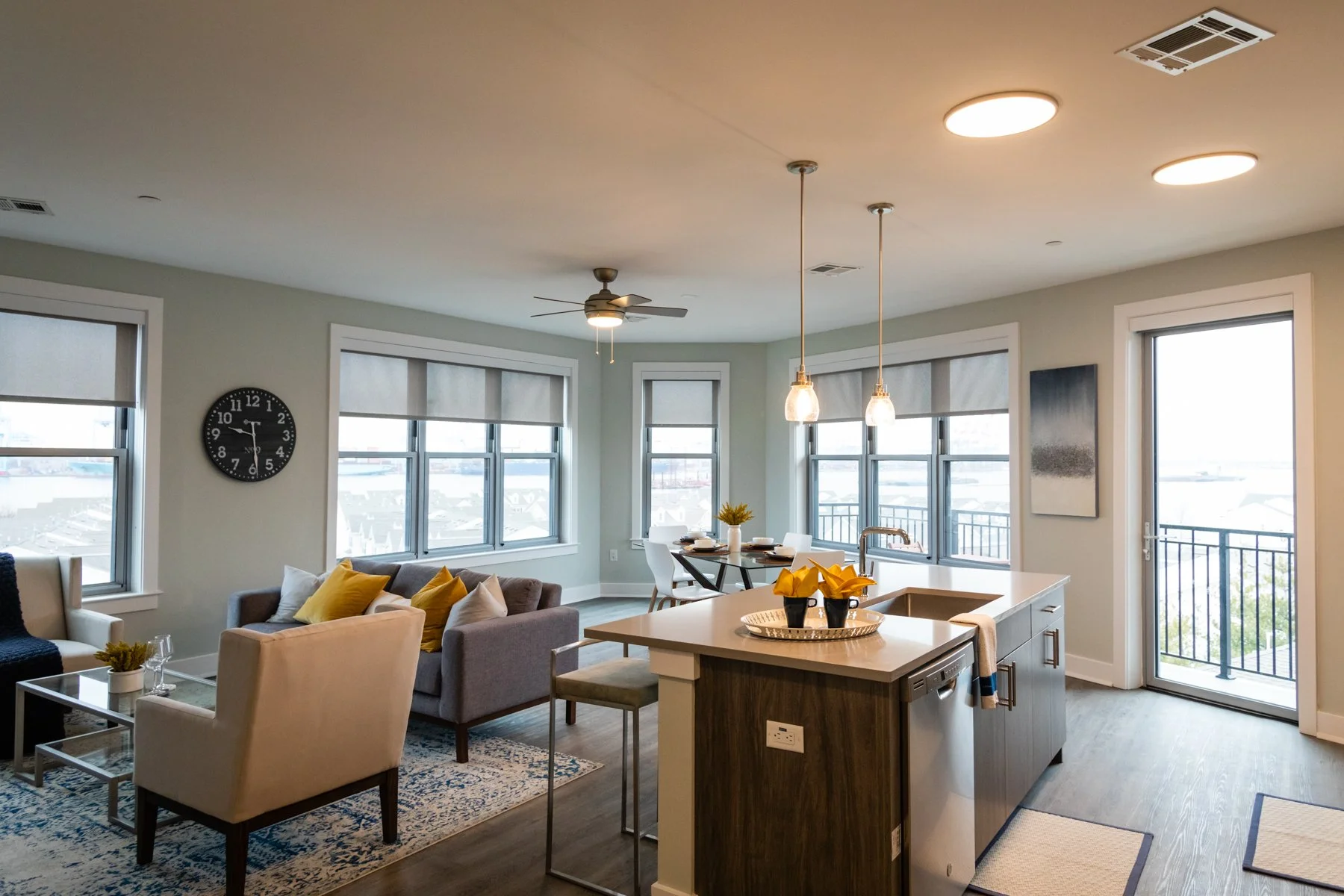 Open-plan living room and kitchen with large windows, a gray sofa with yellow and white cushions, a white armchair, a patterned rug, a round dining table, and a balcony door.