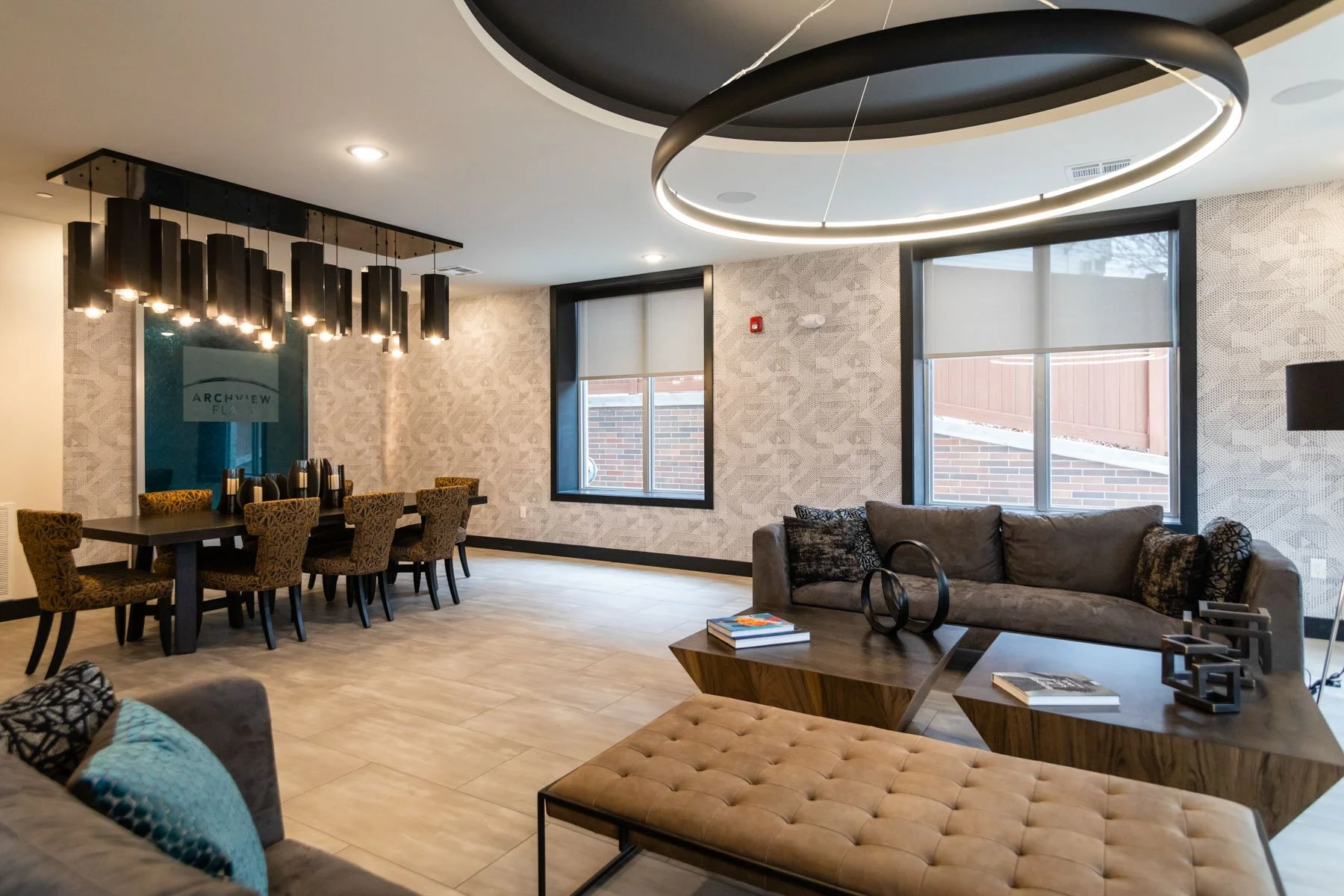 Modern living room and dining area with patterned walls, large windows, chandelier, circular ceiling light, sofas, coffee tables, and decorative items.