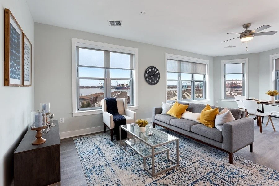 Bright living room with large windows, gray sofa with yellow and white pillows, glass coffee table, white armchair with black cushion, patterned rug, wall clock, and dining area in background.