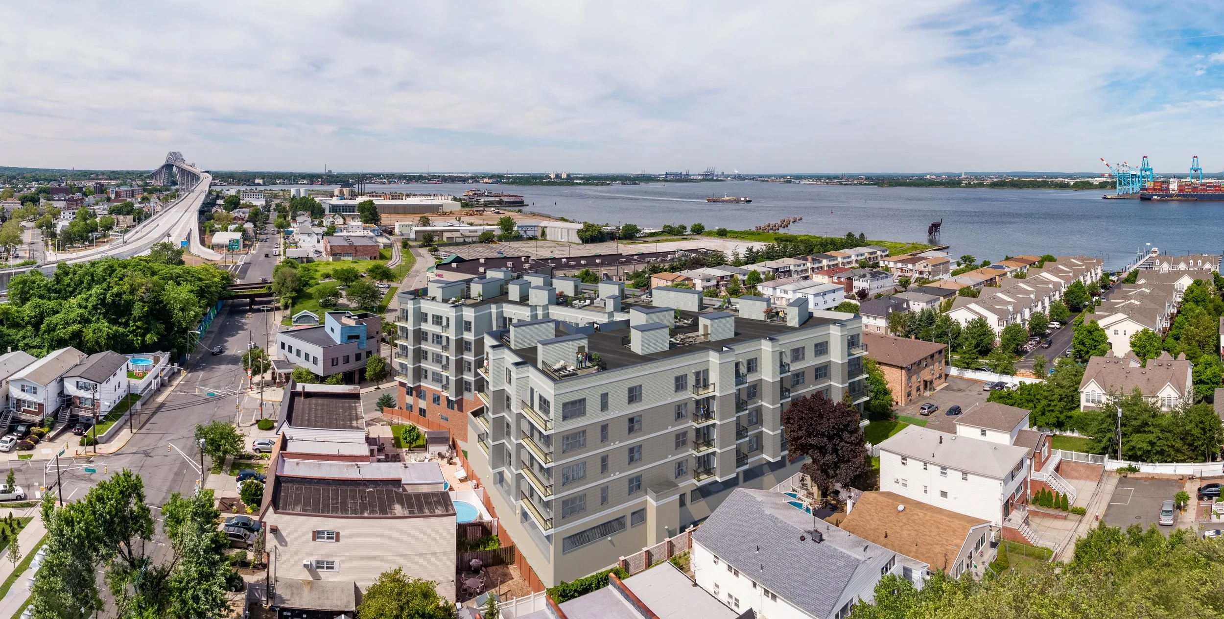 Aerial view of a cityscape near a river, featuring a large bridge, mid-rise apartment building, residential houses, and a harbor with cranes.
