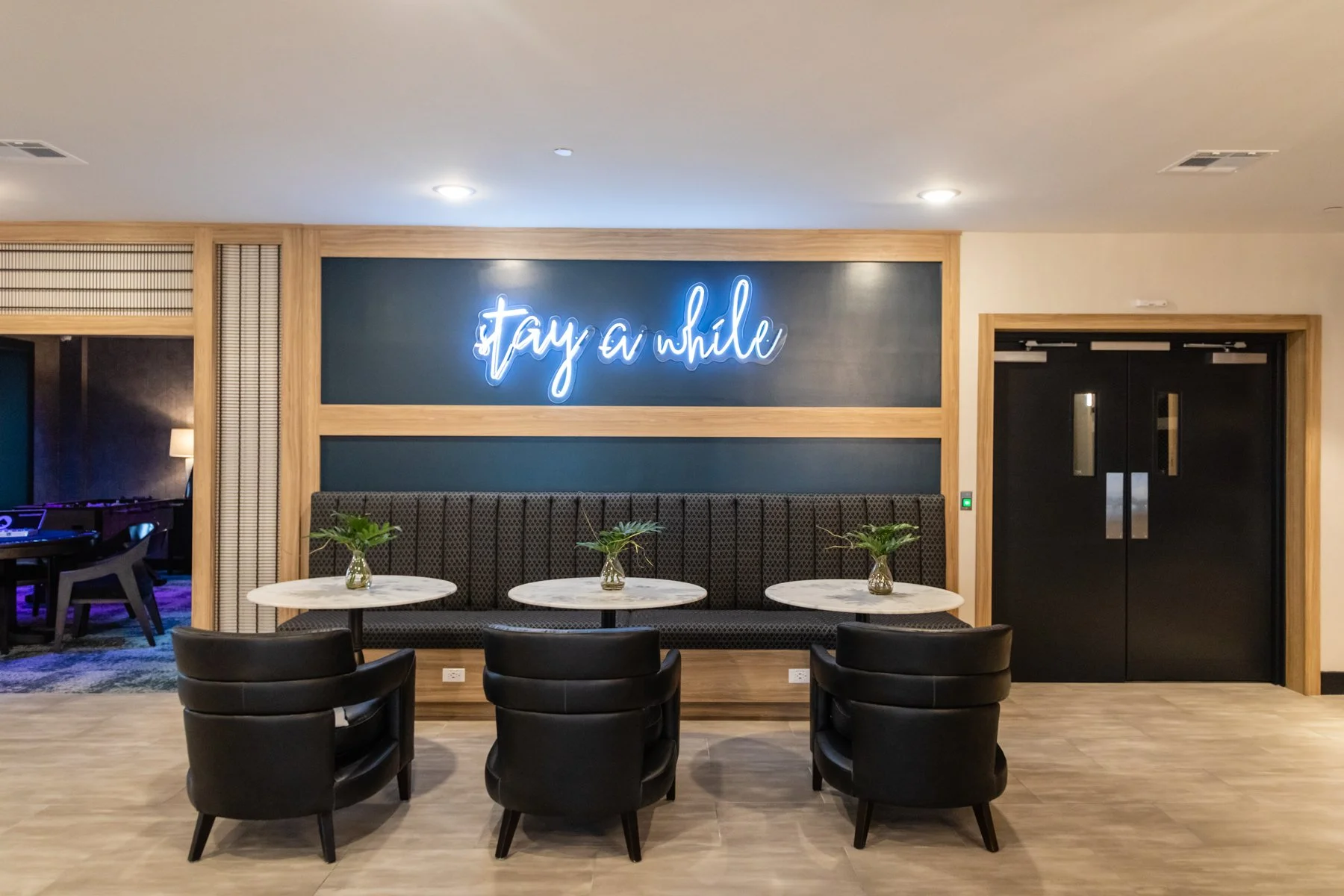 Modern hotel lobby with a gray and black color scheme, featuring a neon sign that says ''stay a while'' on the wall, black chairs, round marble tables with small green plants, and black double doors to the right.