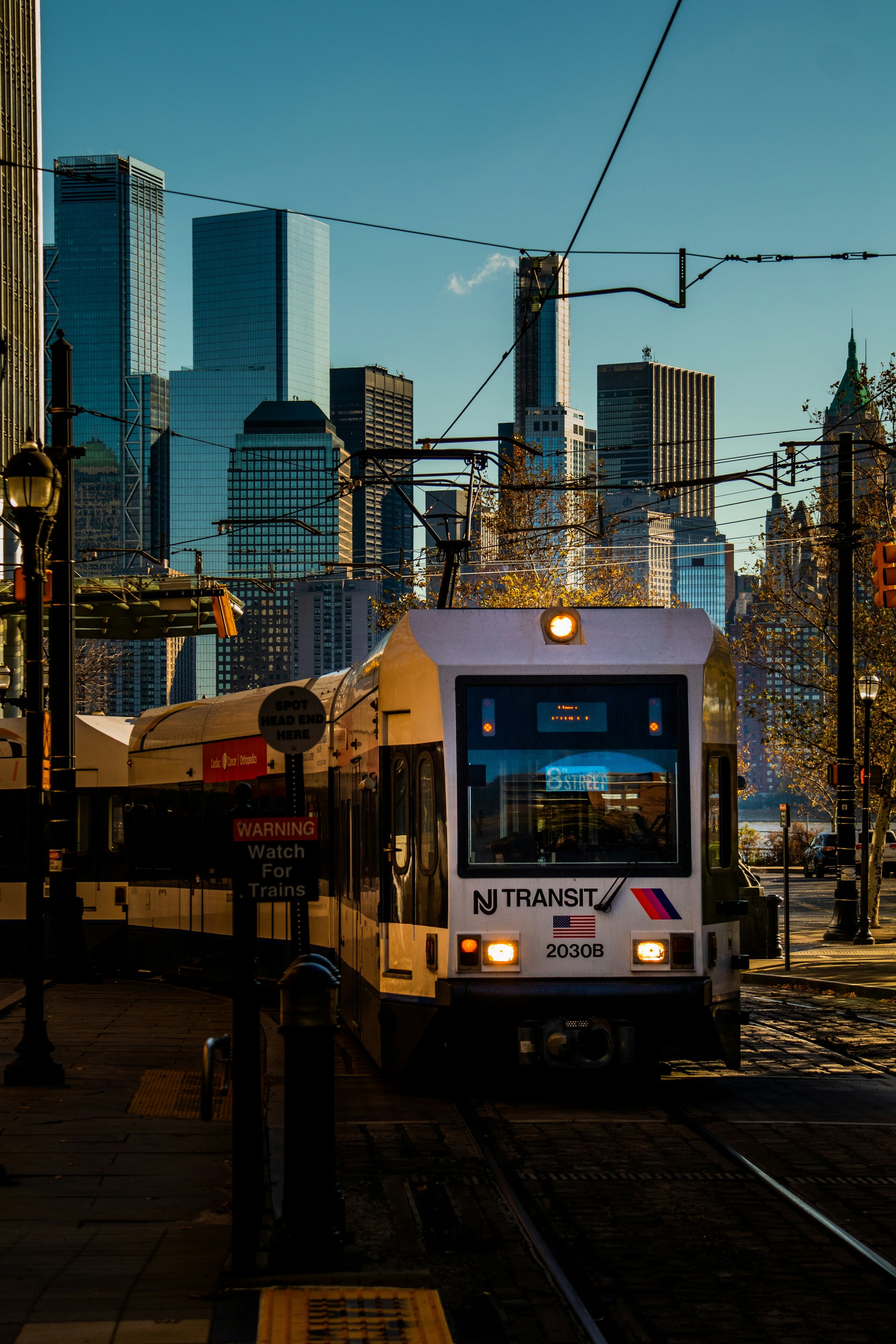 An NJ Transit light rail train on tracks in an urban city with tall skyscrapers in the background, during late afternoon or early evening.