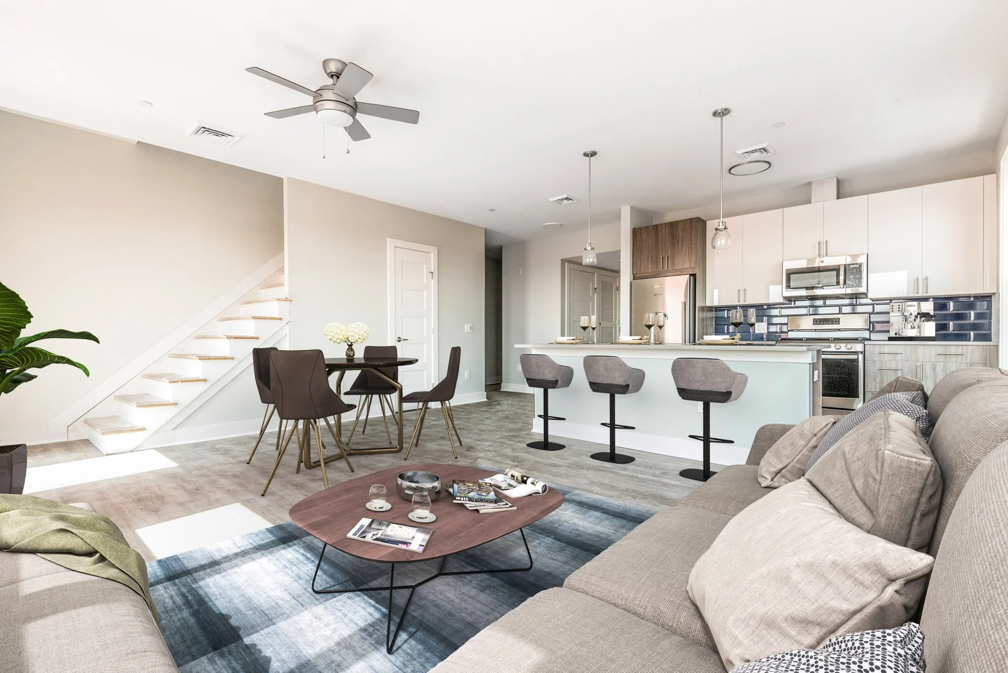 Open-concept living room and kitchen in modern home with gray sofa, wooden coffee table, dining table with six chairs, kitchen bar with three gray stools, stainless steel appliances, gray, white, and black backsplash, and ceiling fan.