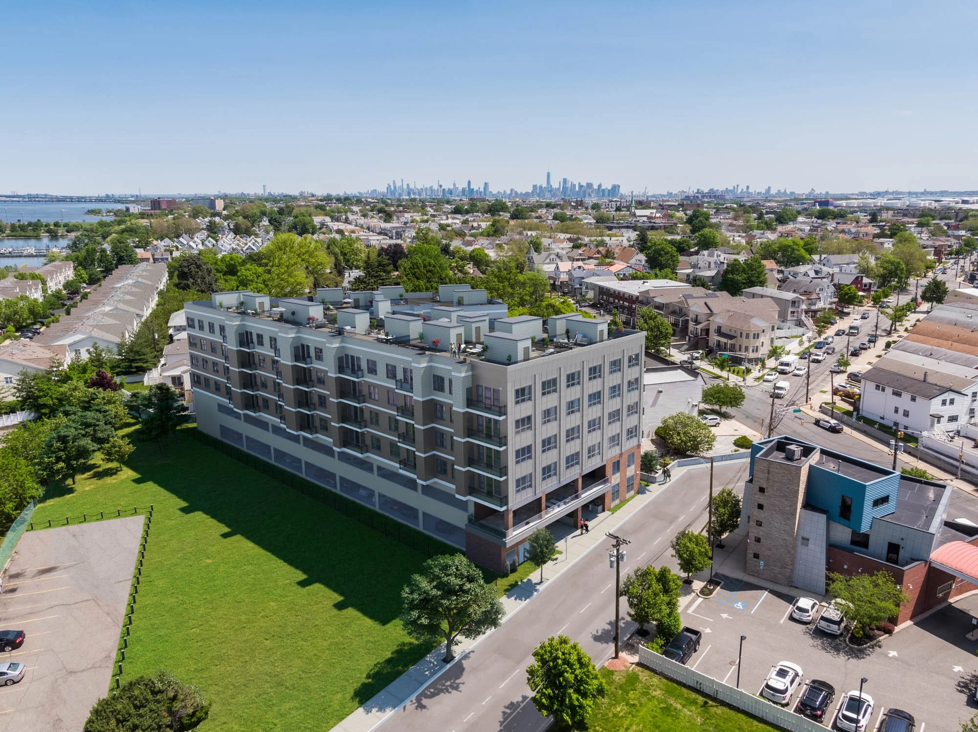 Aerial view of a modern residential apartment building with a green lawn, surrounded by a neighborhood with similar houses, trees, streets, and parked cars, with a city skyline in the background on a clear day.