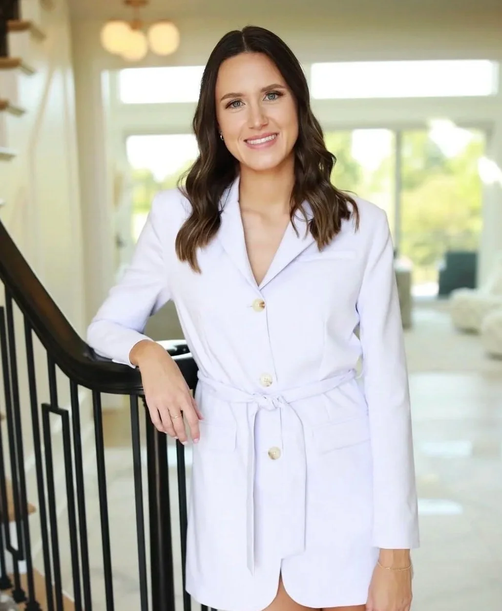Woman with long brown hair wearing a white dress standing beside a staircase railing, smiling in a bright indoor space with large windows.