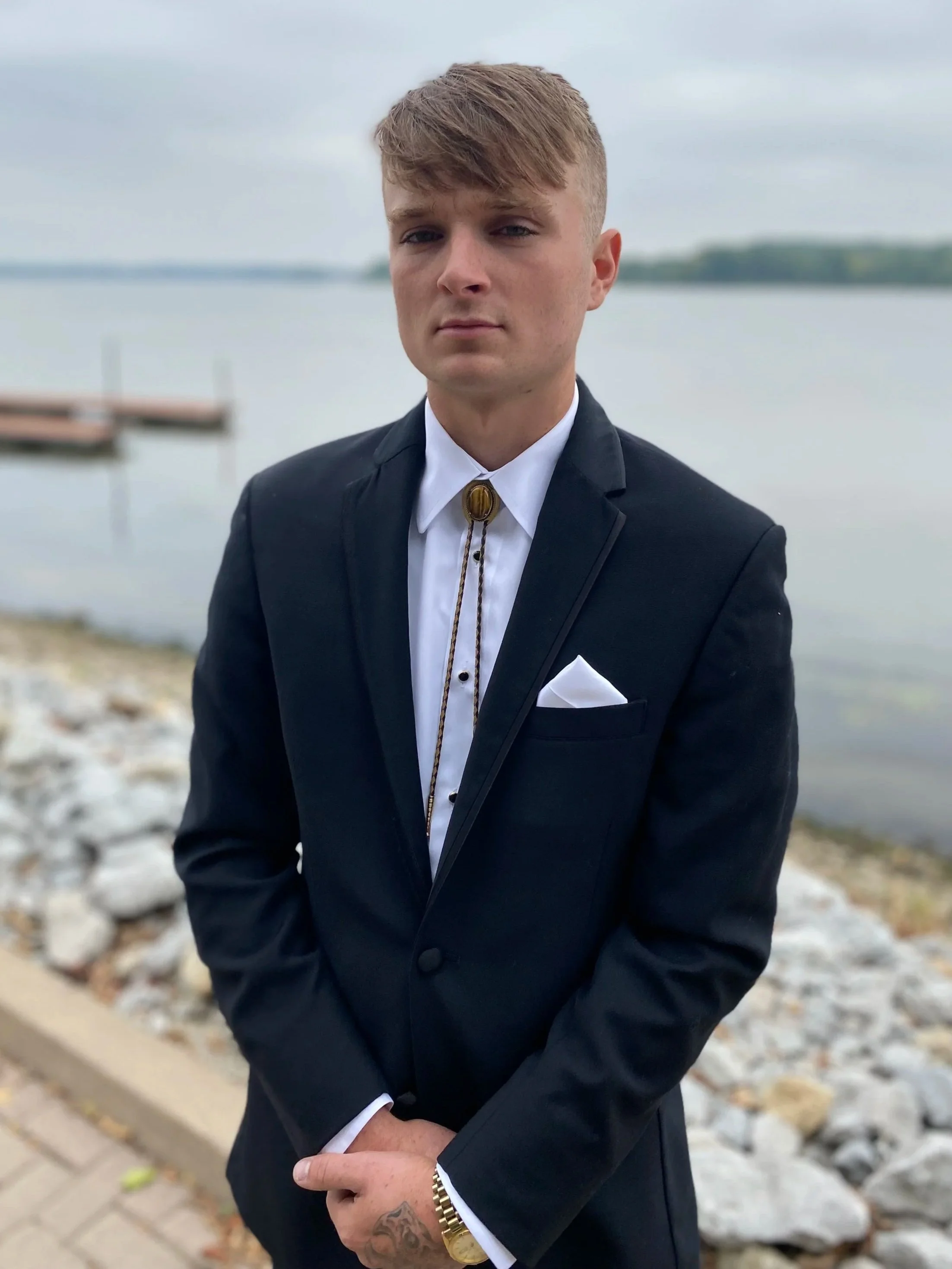 Young man in formal black suit with white shirt, bolo tie, and gold watch, standing outdoors near a lake with rocks and a dock in the background.