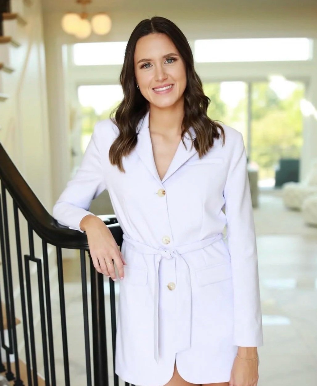 A woman with long dark hair wearing a white dress with gold buttons and a waist tie, standing indoors near a staircase with a railing, smiling at the camera.
