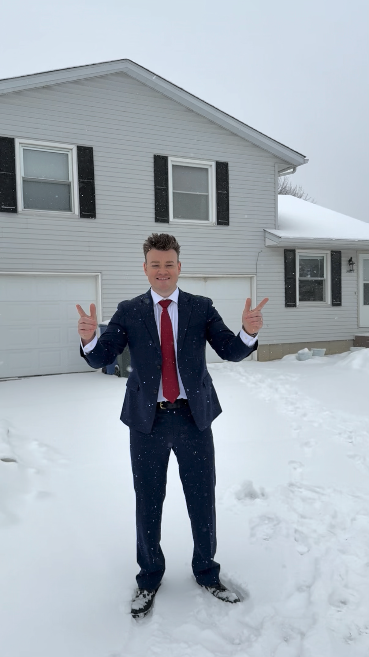 A man in a dark suit with a white shirt and red tie standing outside in the snow in front of a two-story white house with black shutters, smiling and making peace signs with both hands.
