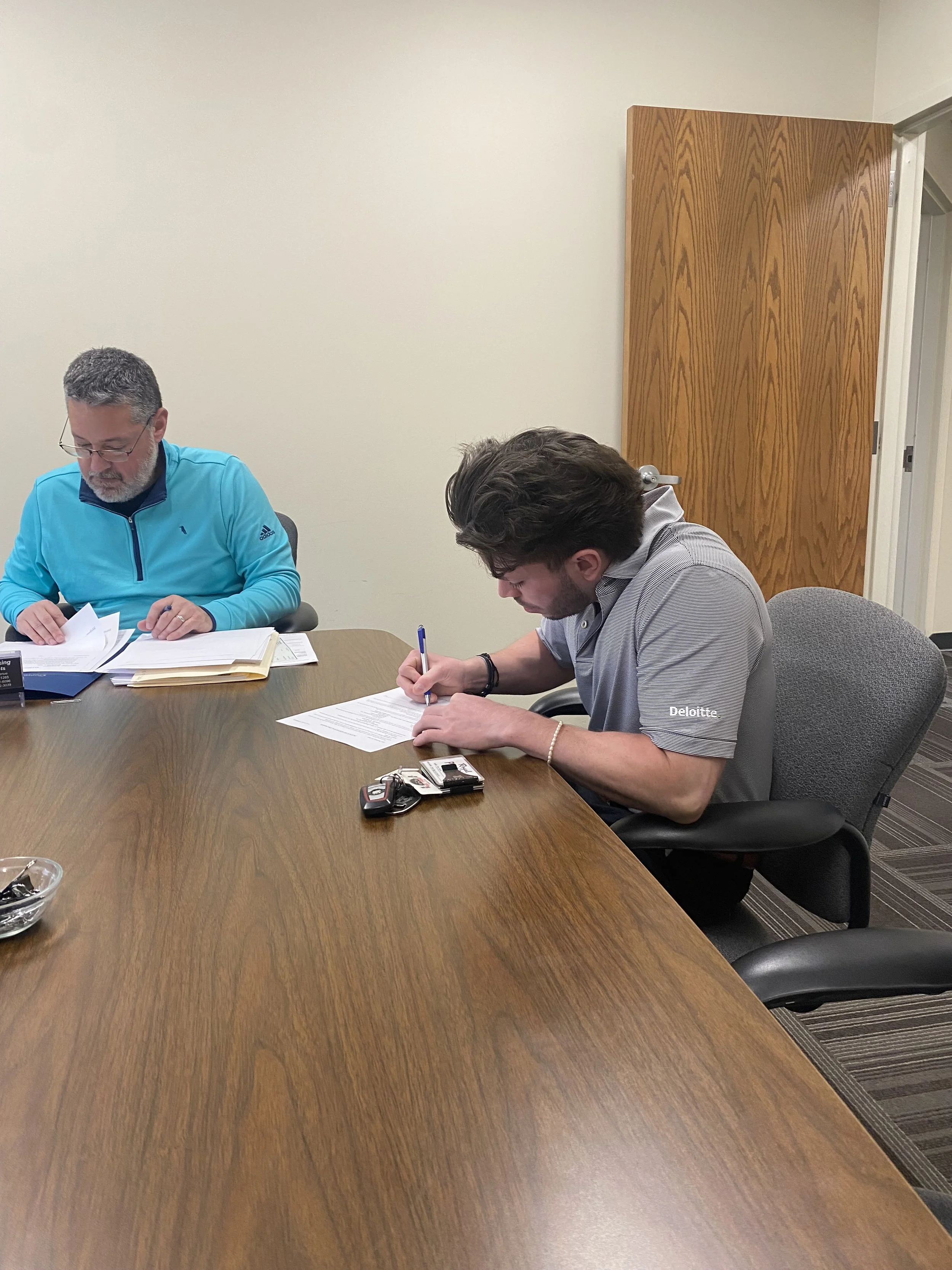 Two men seated at a wooden conference table, reviewing documents. One is wearing a blue jacket, and the other is wearing a gray t-shirt with the Deloitte logo, writing on a paper. There are keys and a small bowl on the table.