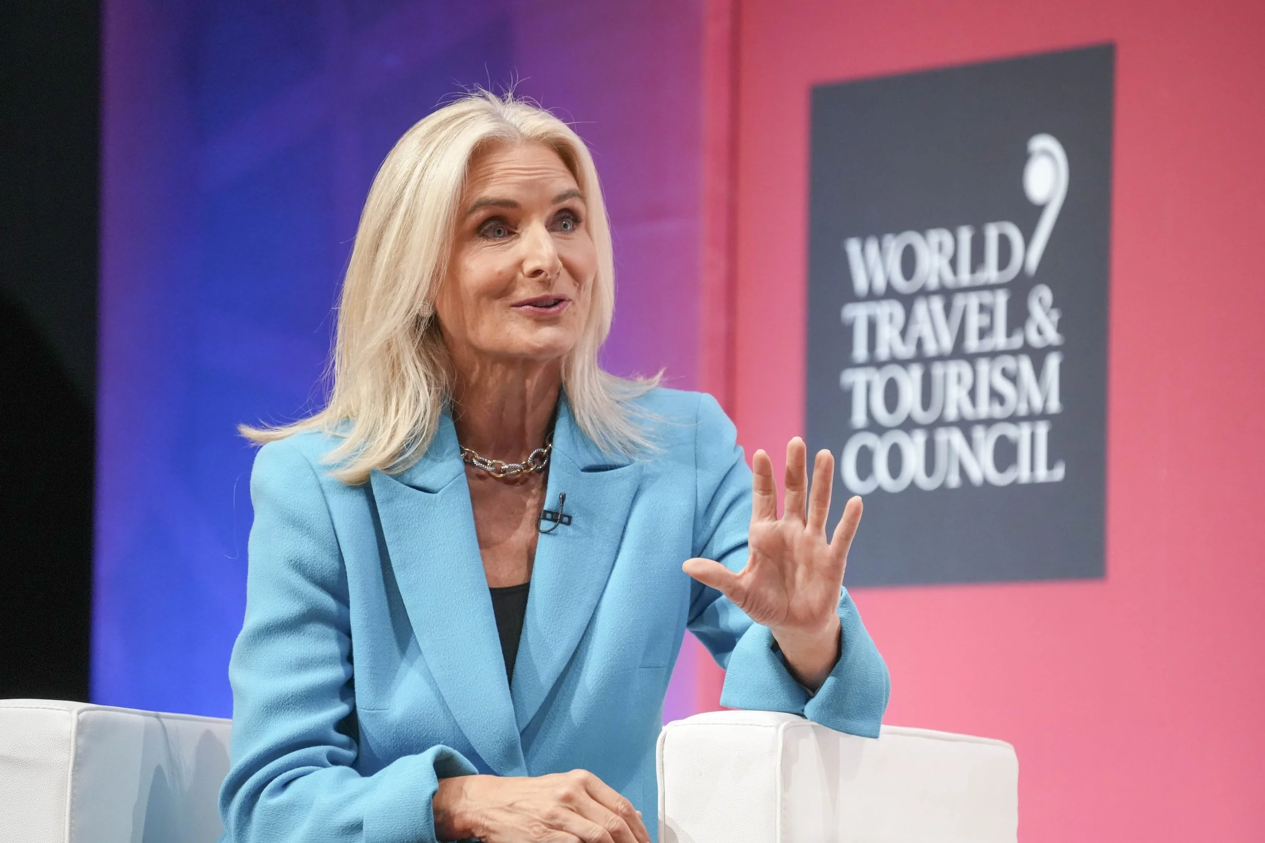 Kelly Craighead wearing a light blue blazer and a silver necklace, speaking at a conference with a sign behind her that says "World Travel & Tourism Council."