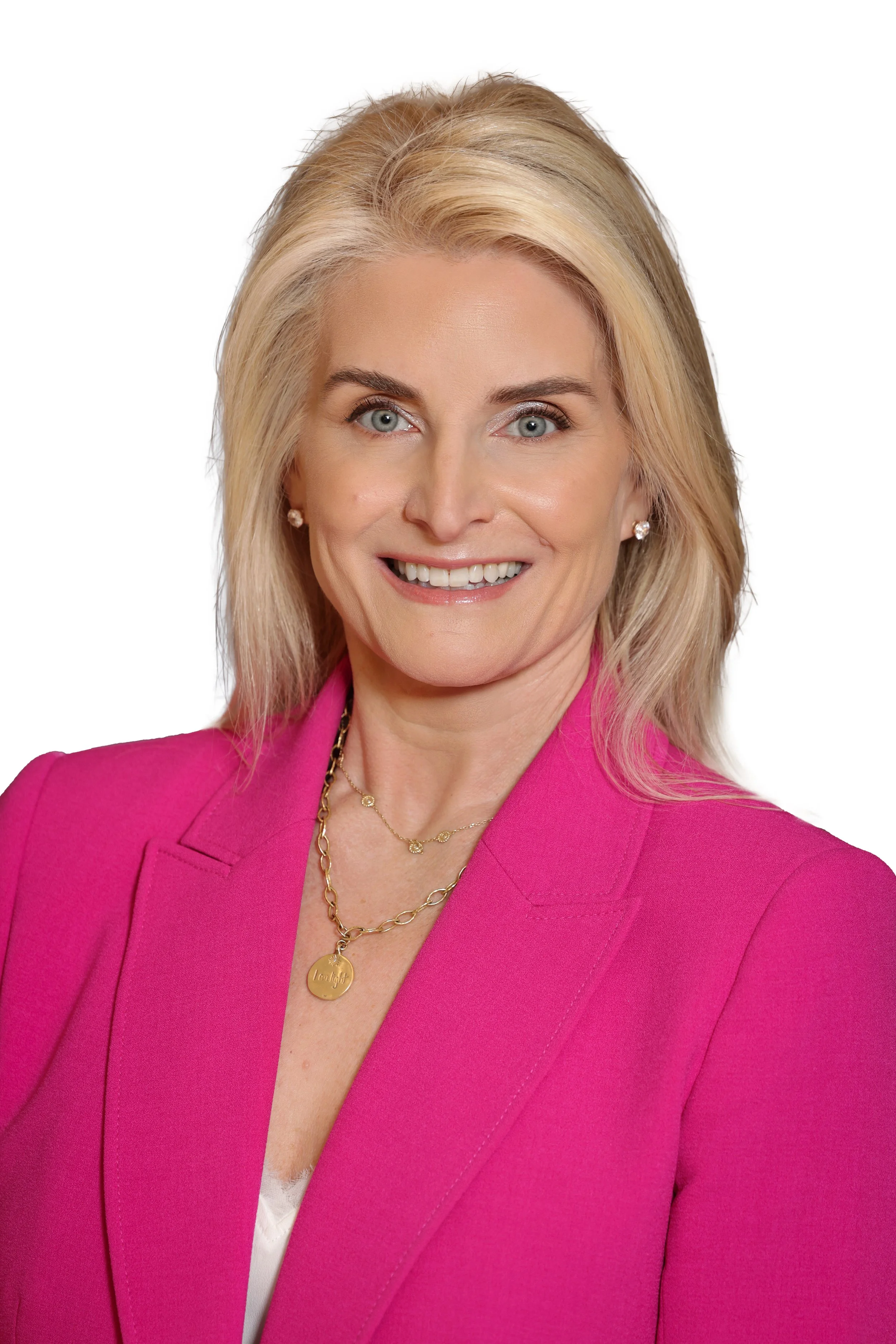 Kelly Craighead wearing a pink blazer, gold jewelry including a necklace with a circular pendant, and small earrings, against a white background.