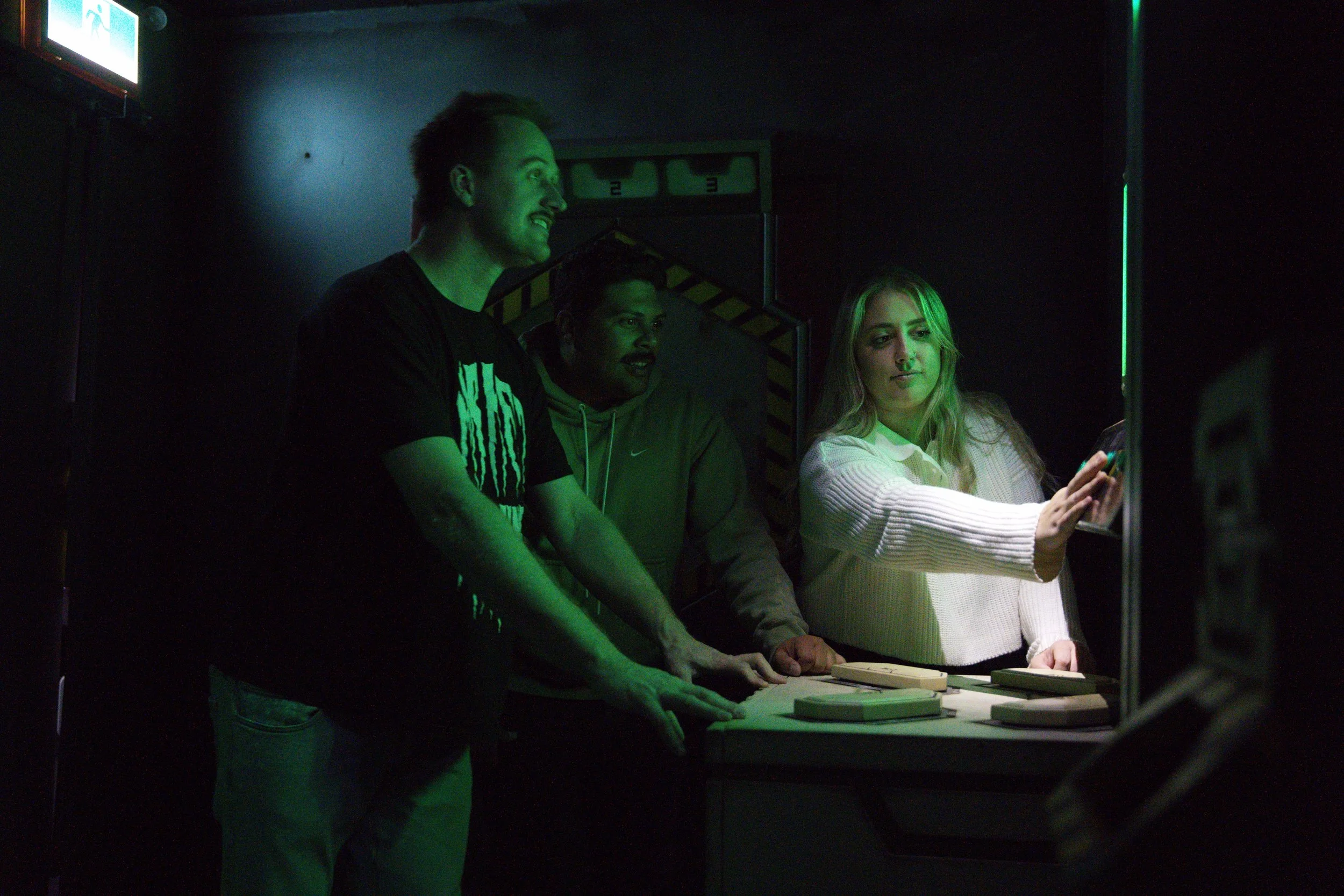 Three young adults at an arcade or gaming venue, with dim lighting and green neon lights, some playing or interacting with a game on a machine.