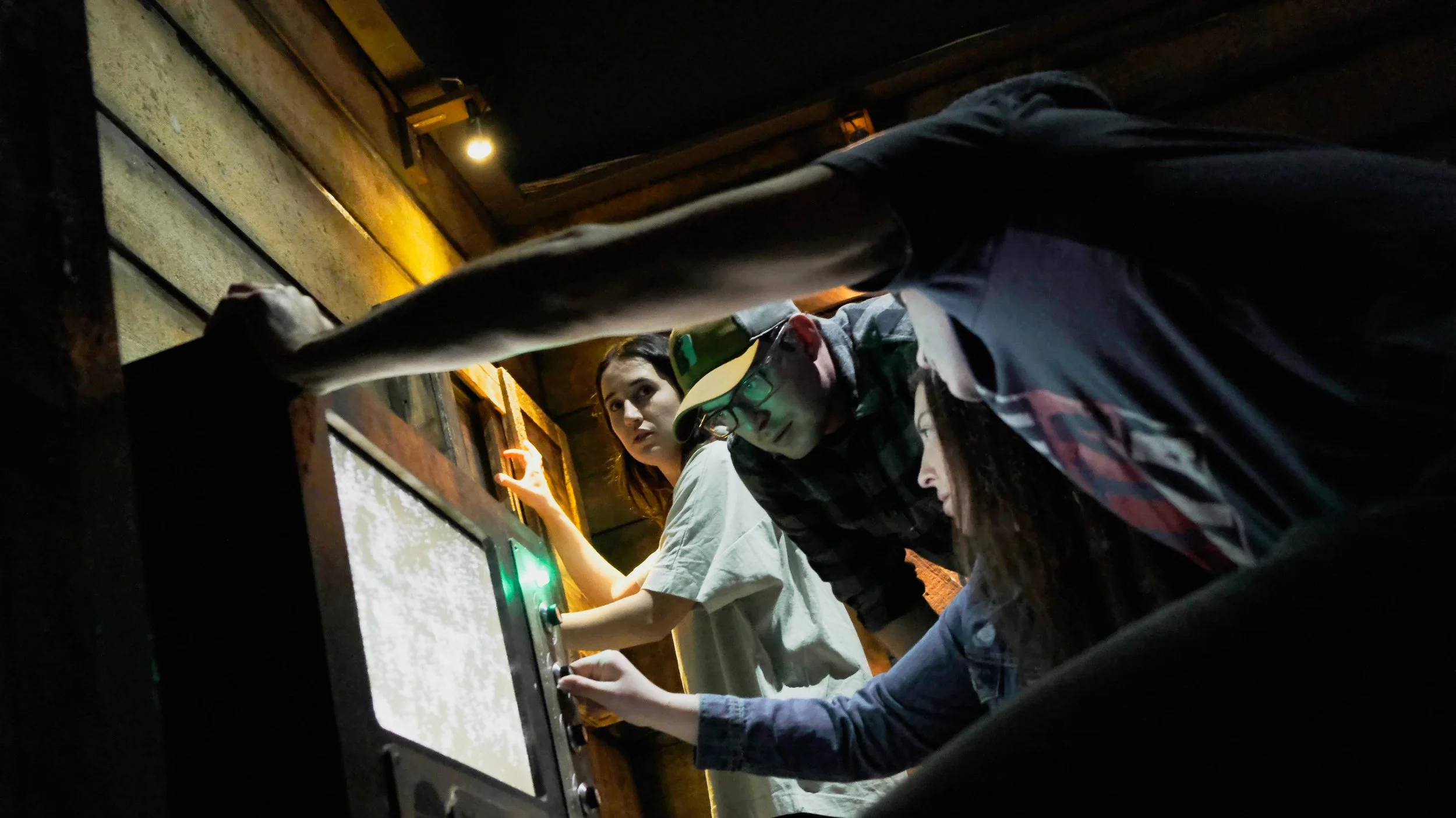 Three people in a dark room operating a control panel, with one wearing glasses and a cap, and the others looking at the panel.