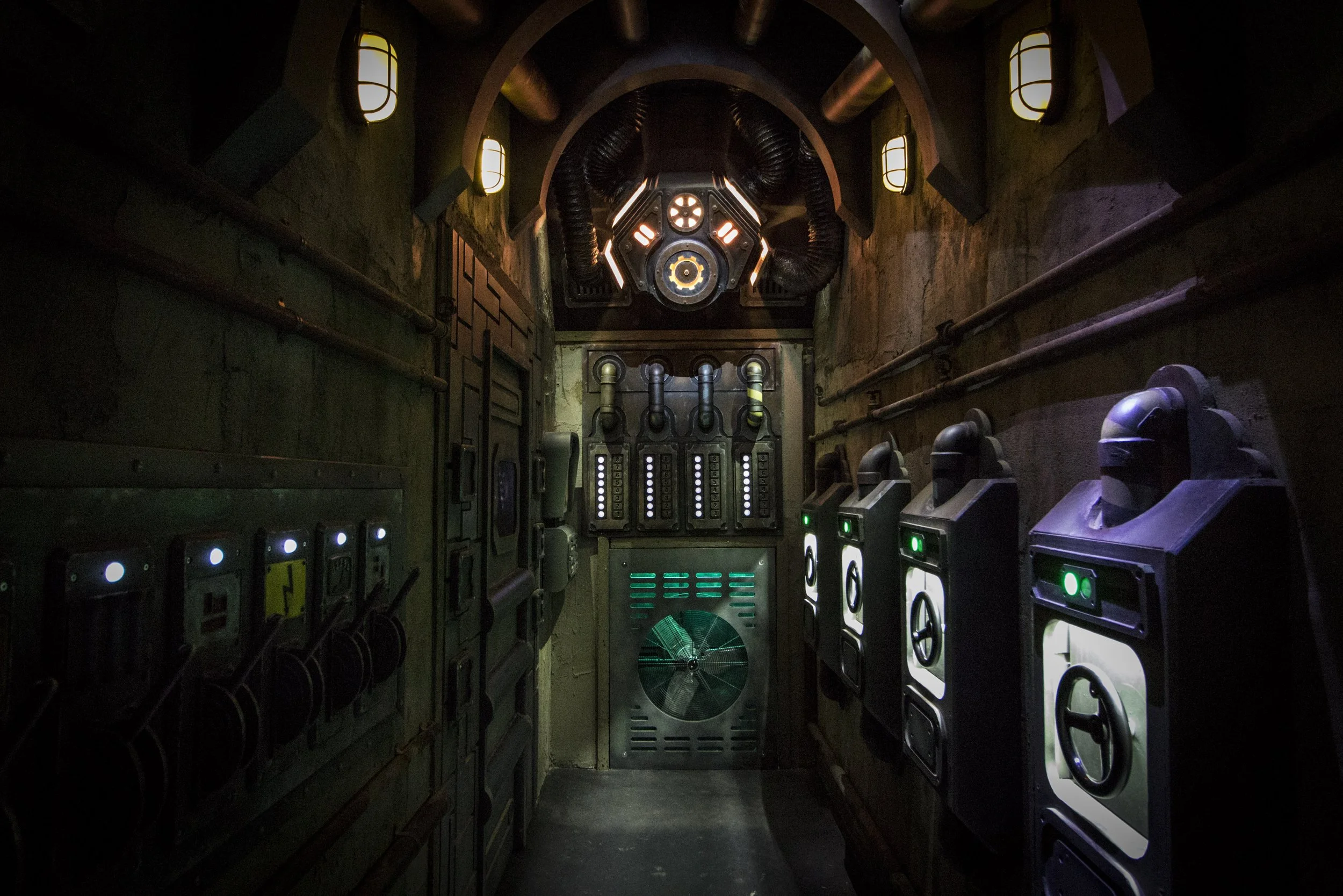 A sci-fi themed corridor with various control panels, buttons, and glowing lights on the walls, leading to a round door at the end. The ceiling has pipes and a central large circular device with vents.