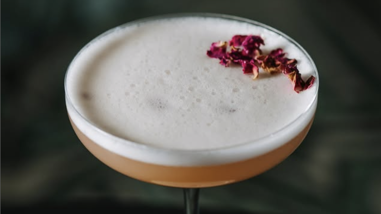 A tall glass with a purple top, filled with a light-colored drink, garnished with green leaves and small white flowers, placed on a black napkin on a wooden surface.