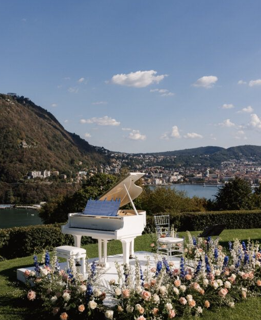 A white grand piano with a blue music sheet stand, surrounded by colorful flowers, outdoors on a lawn with a city and river view in the background, under a partly cloudy sky.