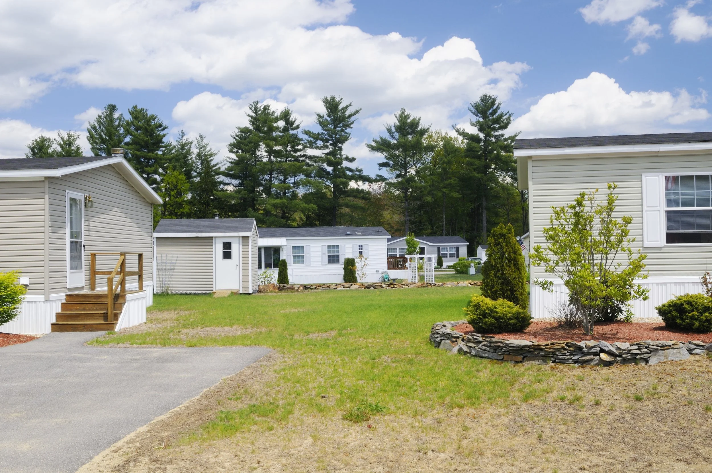 Residential backyard with mobile homes, trees, a small garden, and a partly cloudy sky.