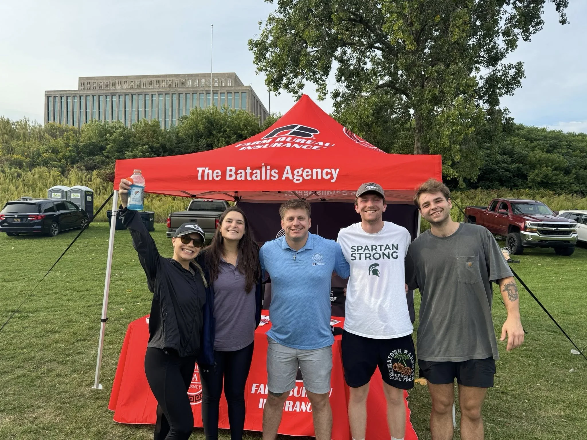 Five young adults standing together outdoors in front of a red tent with the words 'The Batalis Agency' on it. They are smiling and appear happy, with trees and parked cars in the background.