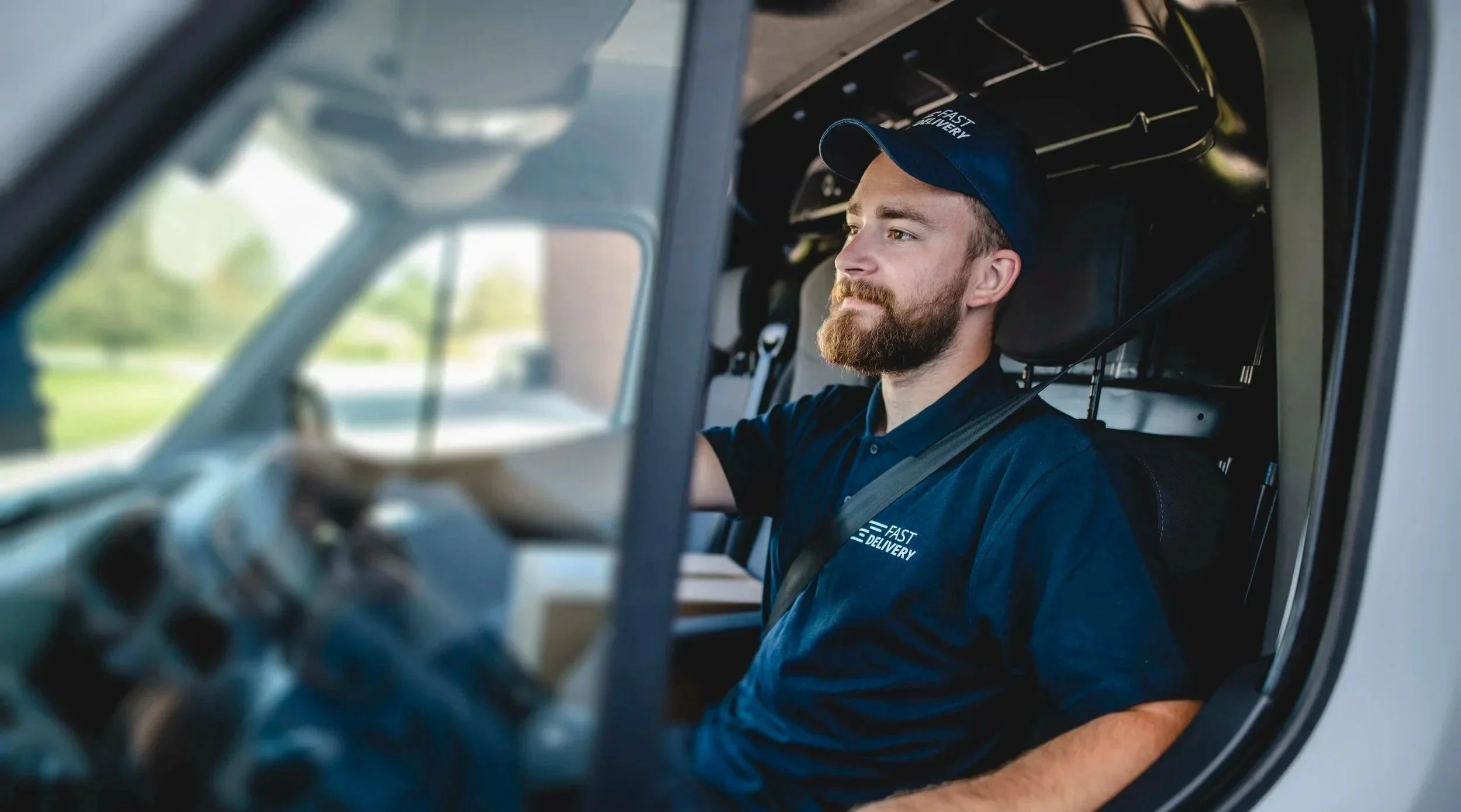 A delivery driver with a beard wearing a navy blue uniform and cap sitting inside a delivery van, looking out the window.
