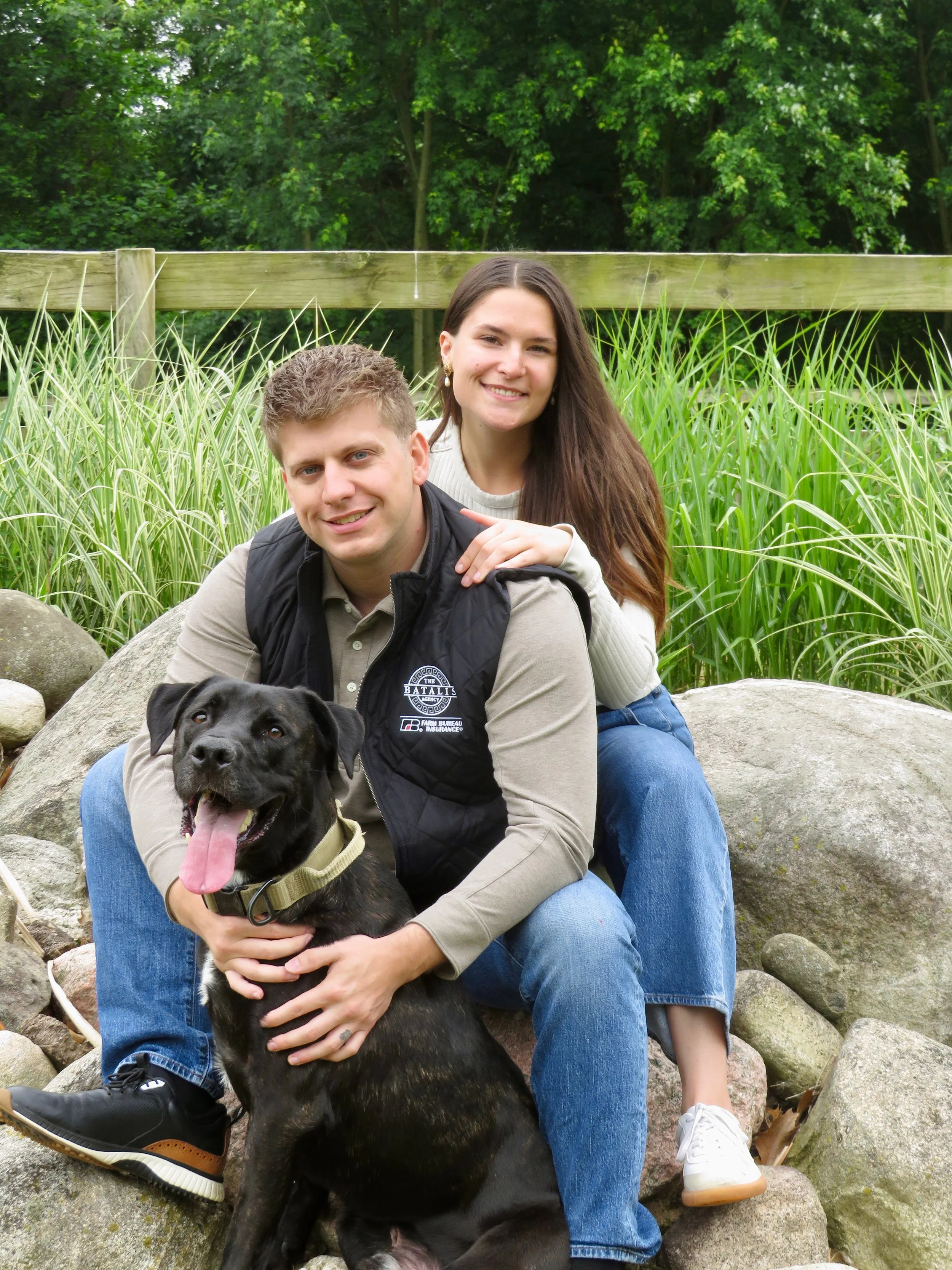 A couple sitting on rocks outdoors with a black dog in front, surrounded by green plants and trees in the background.