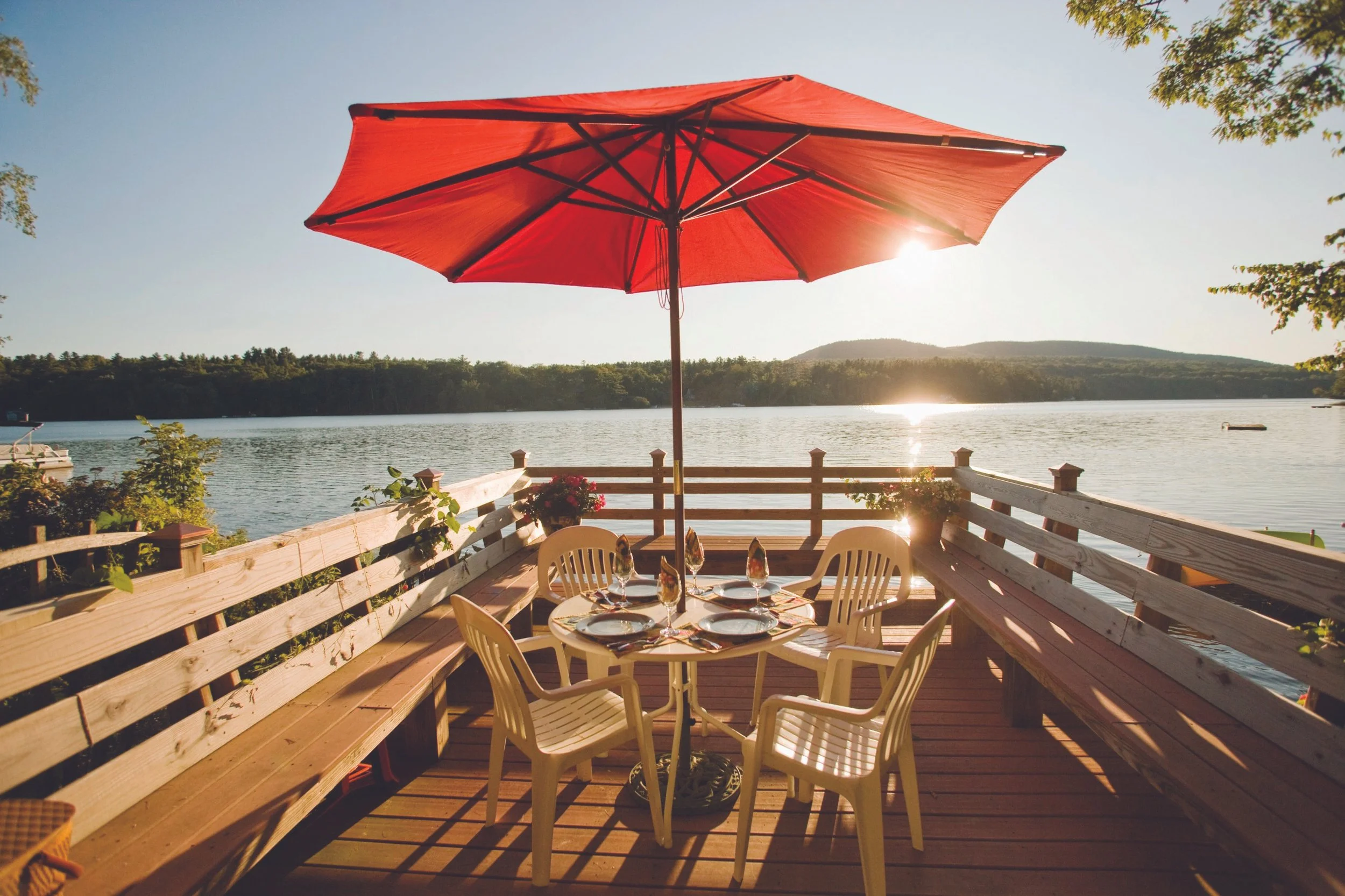 Outdoor dining area on a wooden deck by a lake with a red umbrella, table set with plates and wine glasses, surrounded by a bench and chairs, scenic water and mountains in the background