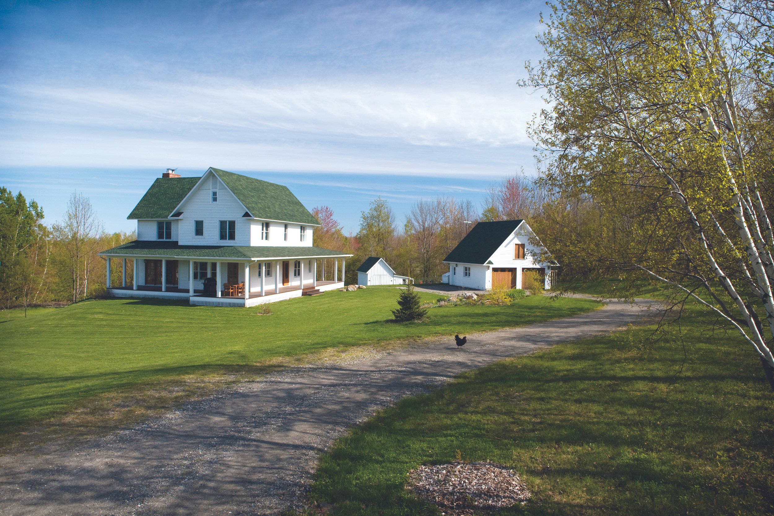 A large white house with a green roof and a wraparound porch, surrounded by a lawn, trees, and smaller white outbuildings, under a partly cloudy sky.
