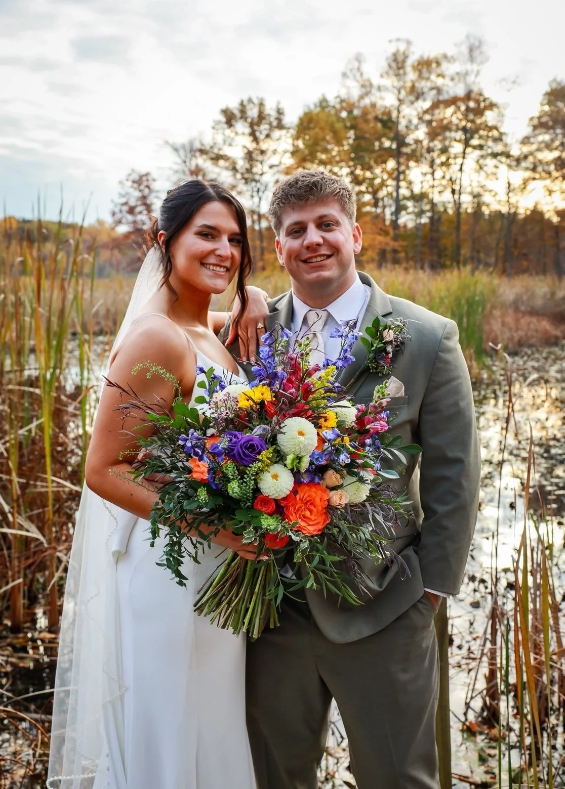 A bride and groom smiling outdoors during fall, holding a large colorful bouquet of flowers, with trees and a pond in the background.