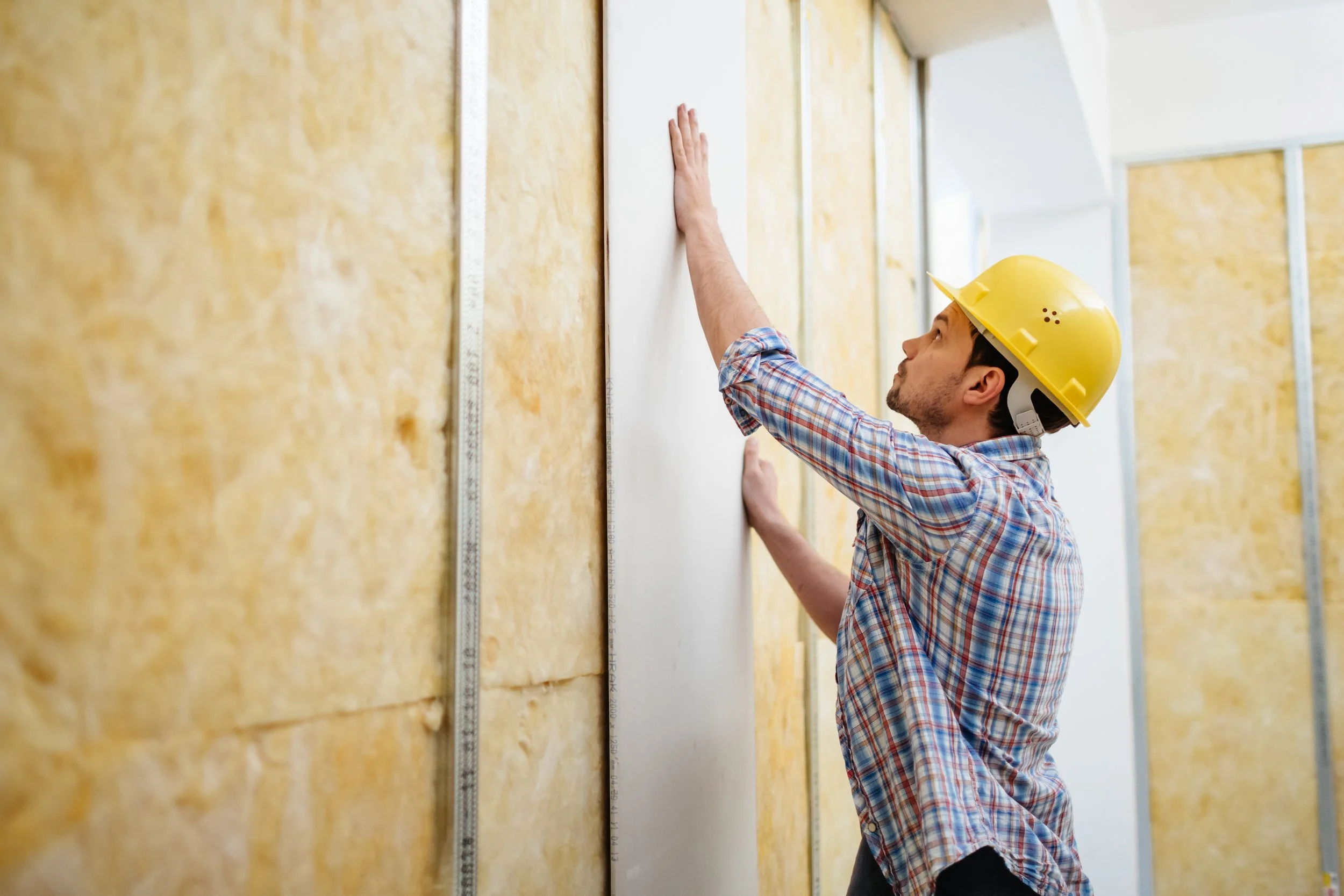 A construction worker wearing a yellow hard hat and plaid shirt installing insulation on a wall.
