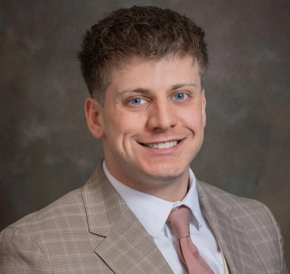 Headshot of a young man with brown hair and blue eyes, smiling, wearing a beige plaid suit, white shirt, and light pink tie, against a dark textured background.