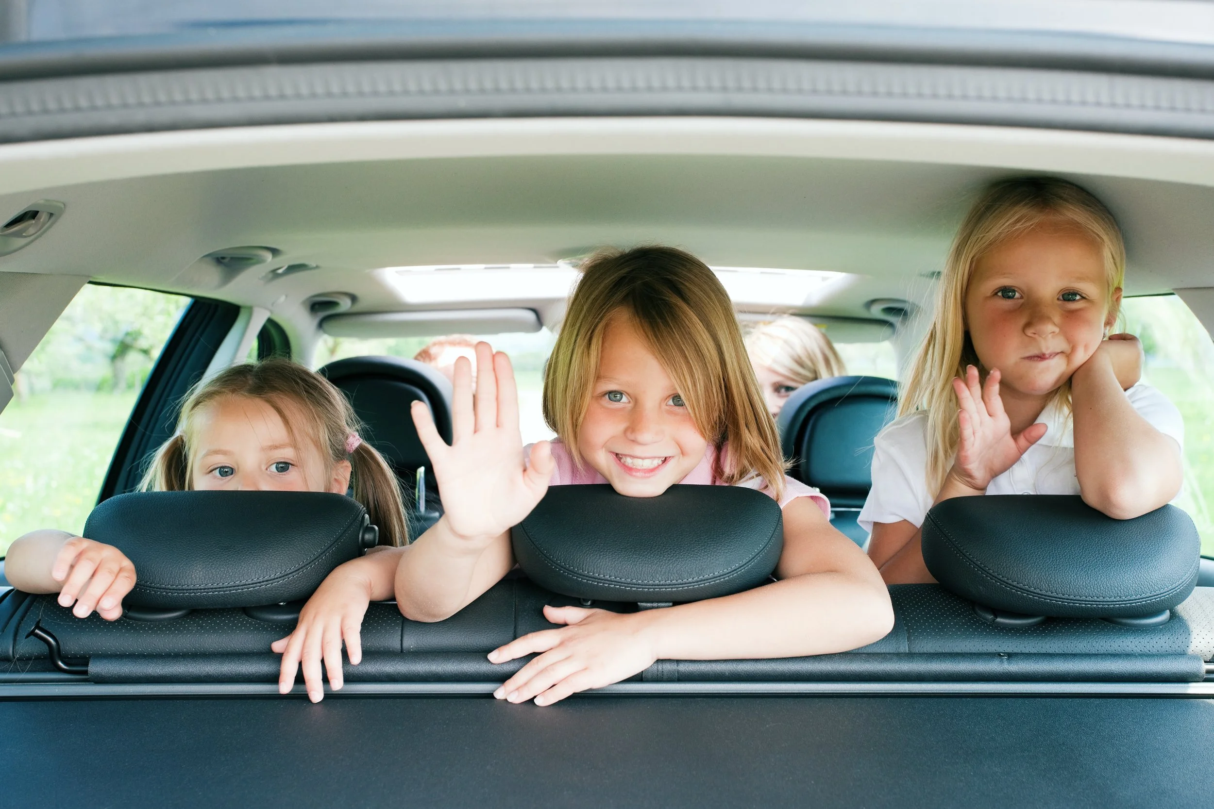 Three young girls are leaning on the back of car seats, looking out through the rear window of a vehicle, with trees and greenery outside.