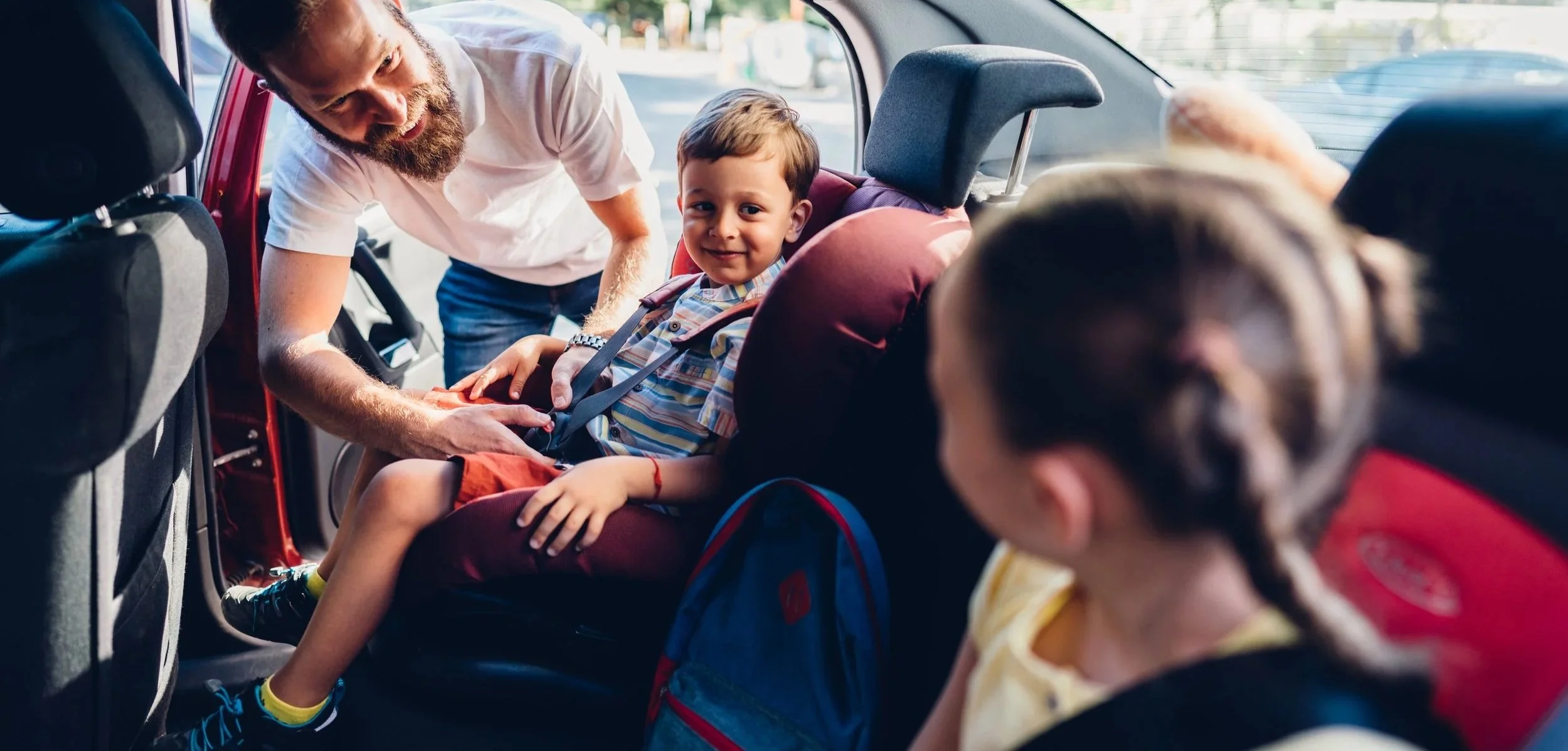 A man helping a young boy secure his seatbelt in a car, with a girl in a car seat in the foreground. The boy is smiling and looking at the camera.
