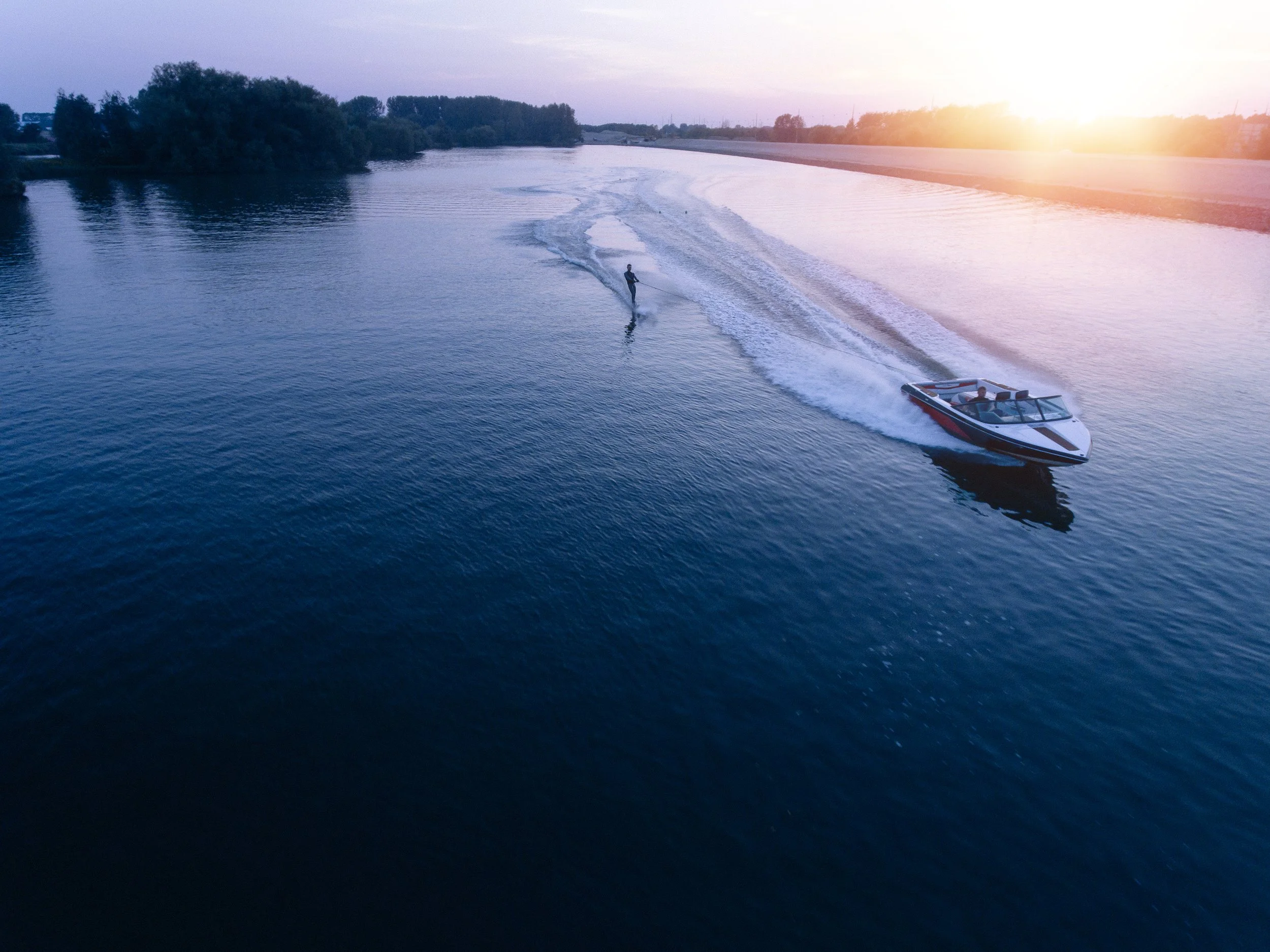 A boat is being driven on a river during sunset, creating waves behind it. There is a person on a water ski being pulled by the boat.
