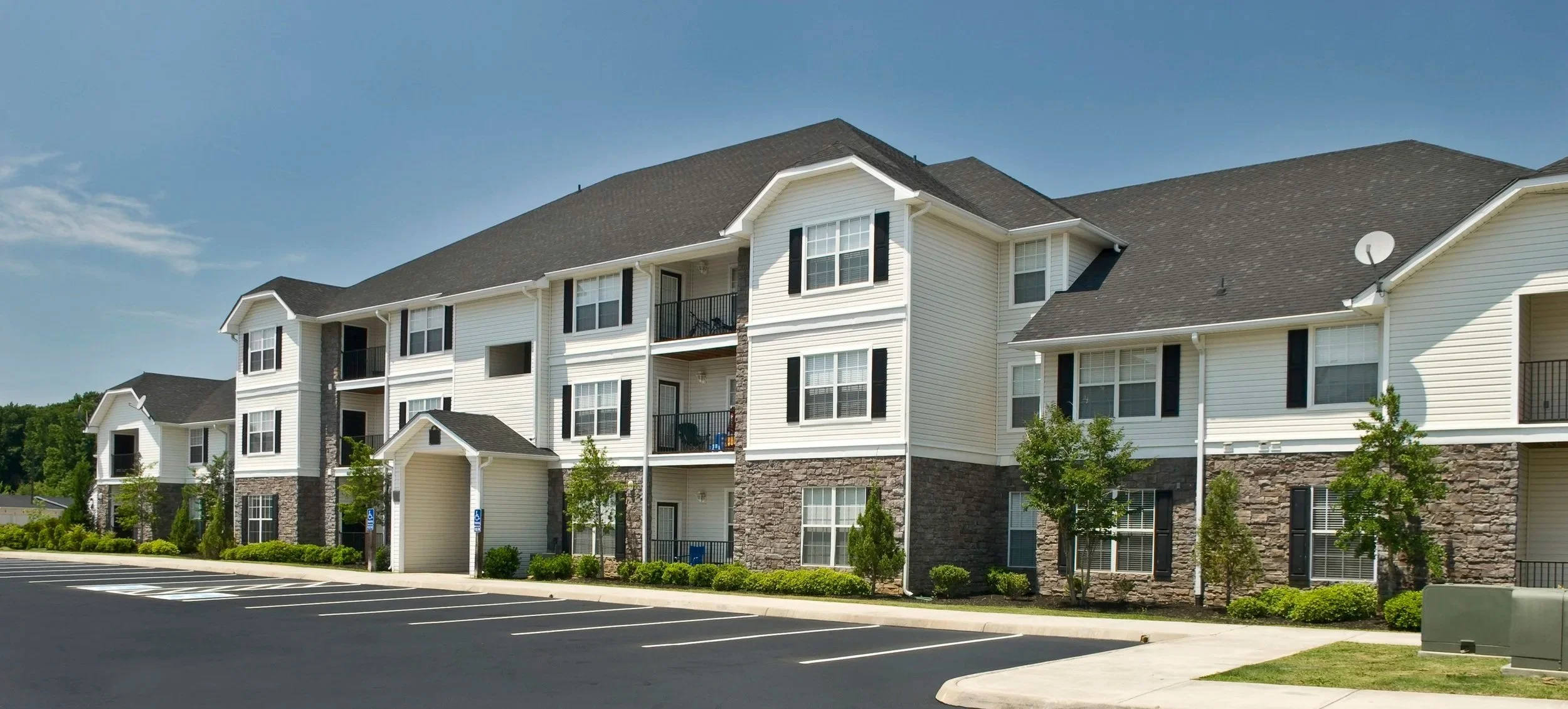 Multi-story apartment building with white siding, black window shutters, stone accents at the base, and small balconies, adjacent to a parking lot with marked parking spaces.