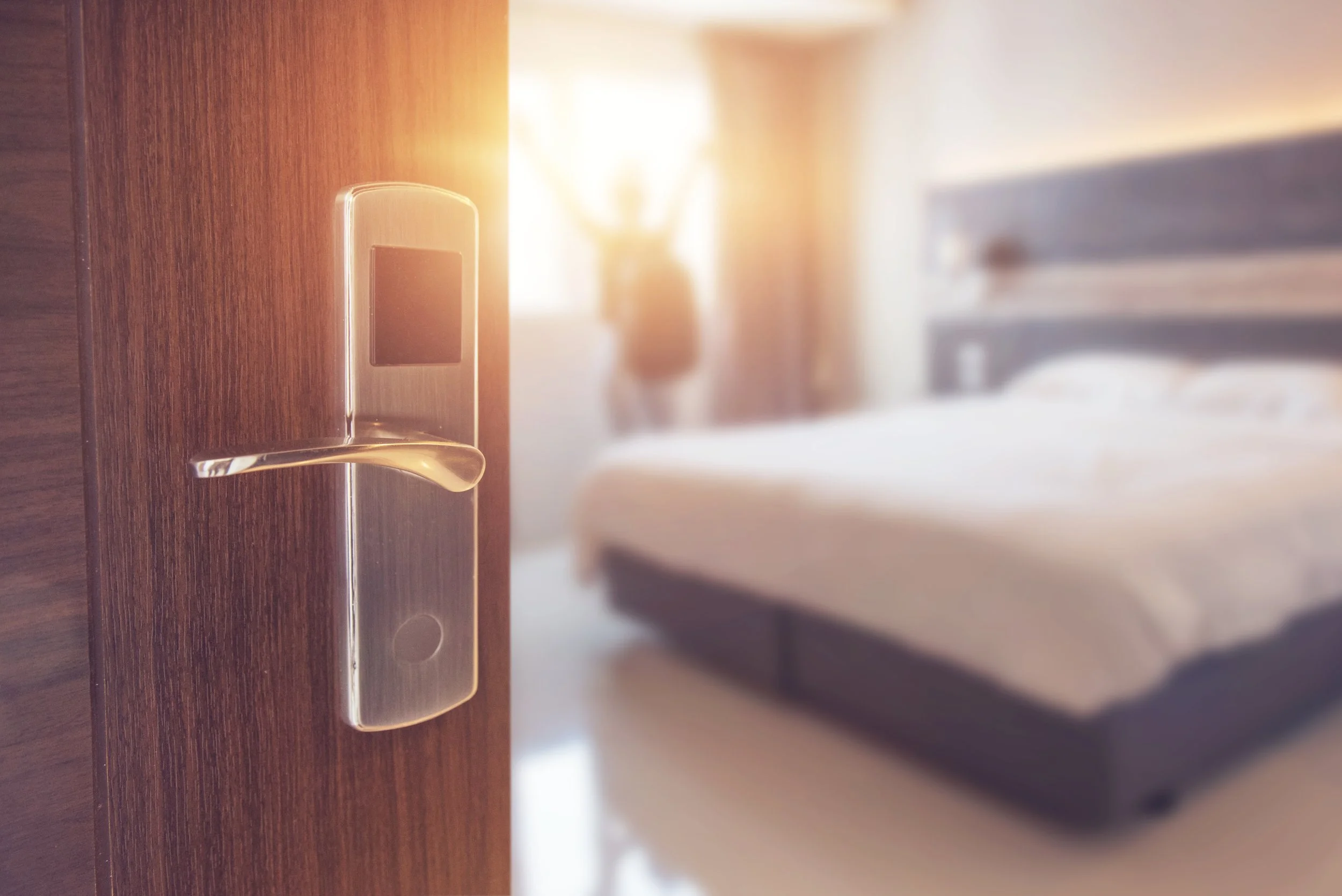 Close-up of a modern digital hotel room door lock with a blurred hotel bed and person in the background.