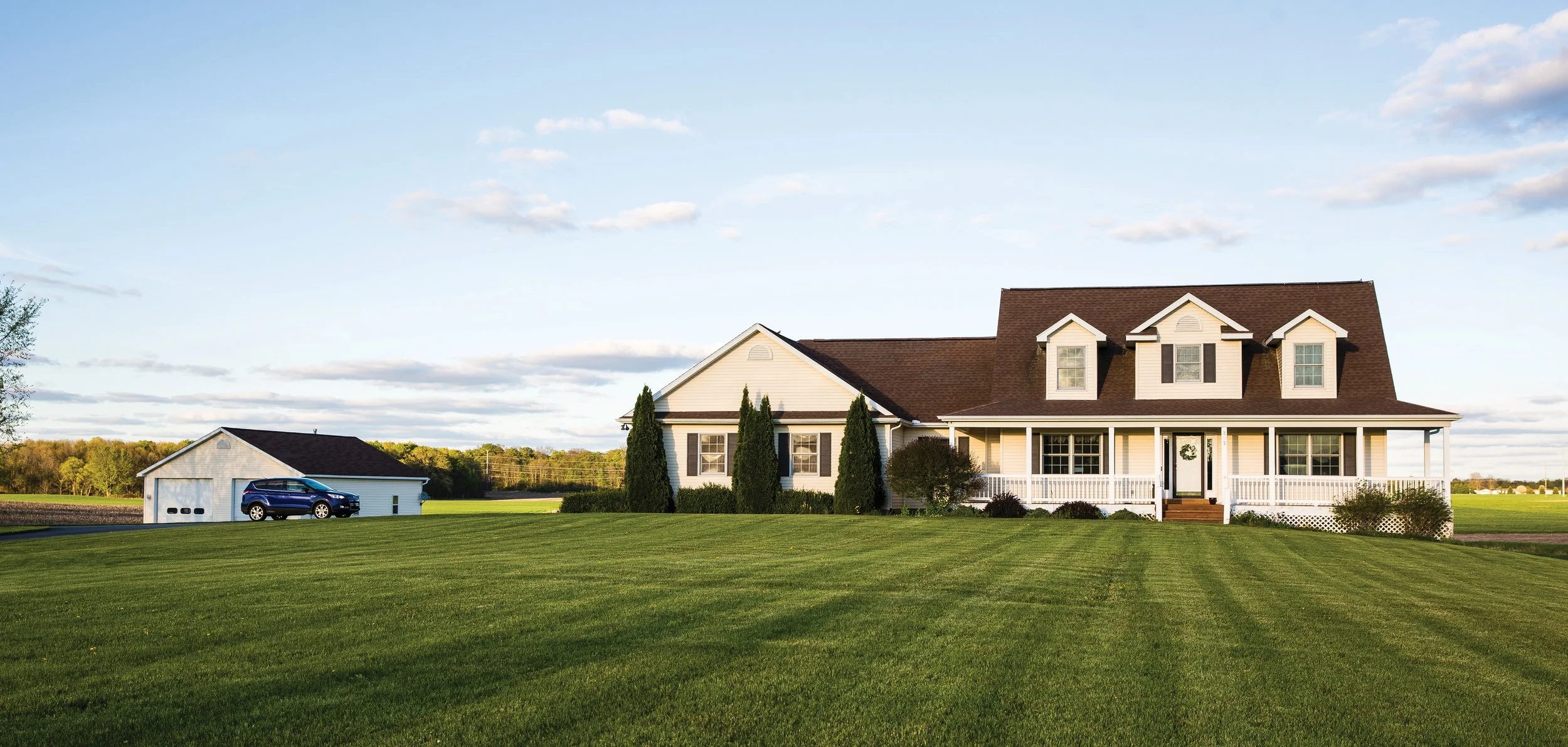 A house with a large front porch, white exterior, and three dormer windows on the roof, surrounded by a well-maintained lawn and a detached garage with a parked vehicle.