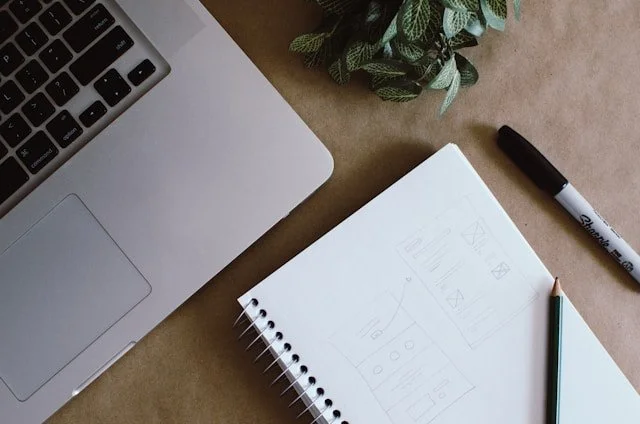 ノートパソコン、白い紙、ペン、マーカーペン、鉛筆、観葉植物が机の上に配置されているデスクワークの様子