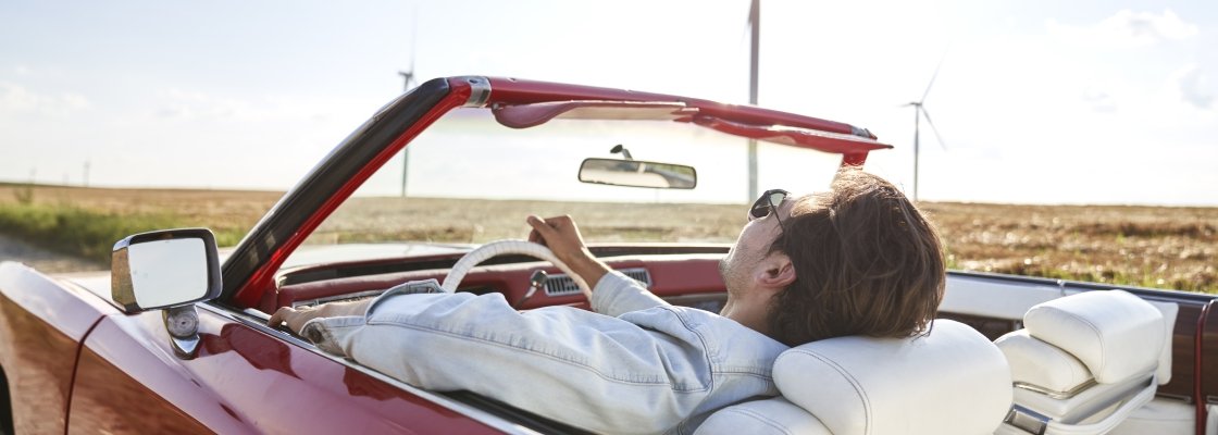 A man lies back in a red convertible car with his eyes closed, enjoying a drive through an open rural landscape with wind turbines in the background.