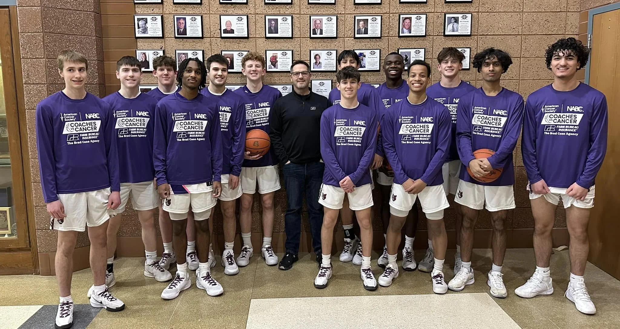 A group of young male basketball players in purple jerseys and white shorts standing in a hallway, posing for a team photo with their coach in the middle. The background features framed photographs on the wall.