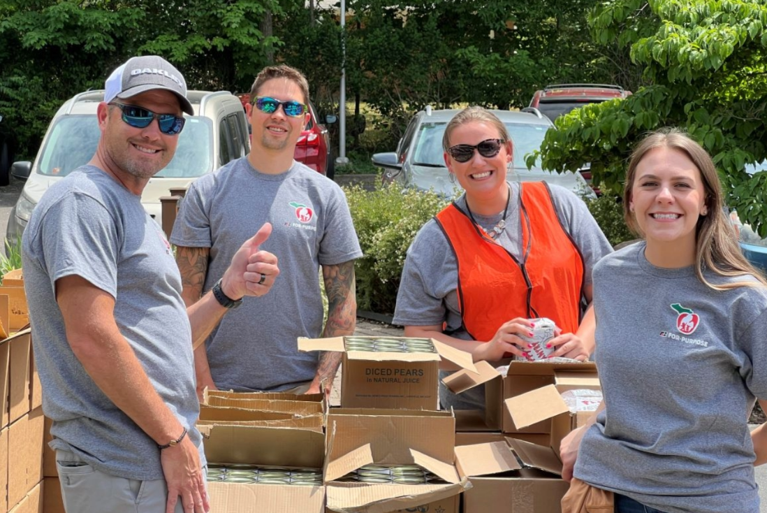 Four people smiling outdoors near boxes of canned goods, two men and two women, with trees and parked cars in the background.