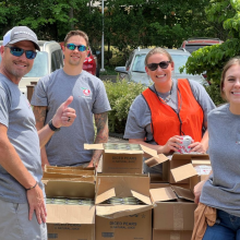 Four people standing outdoors near boxes, smiling and giving a thumbs-up, wearing casual clothes including gray t-shirts and one in an orange vest.
