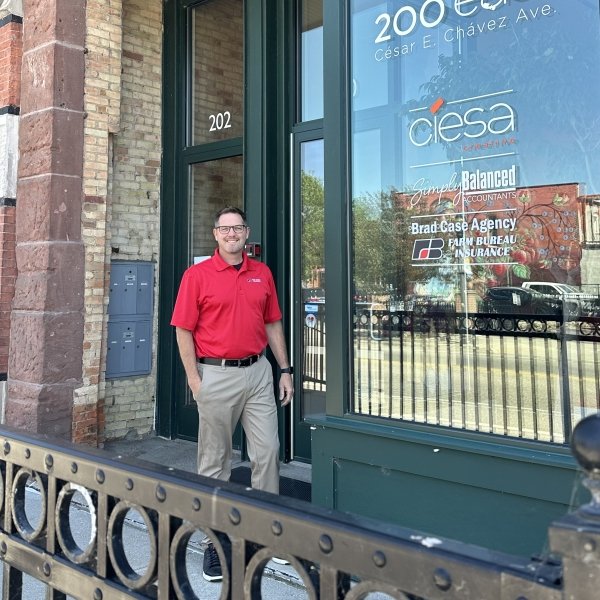 A man wearing a red polo shirt and khaki pants standing outside a building with glass windows and a green frame, next to a black fence and brick wall. Reflections of trees and cars are visible in the windows.