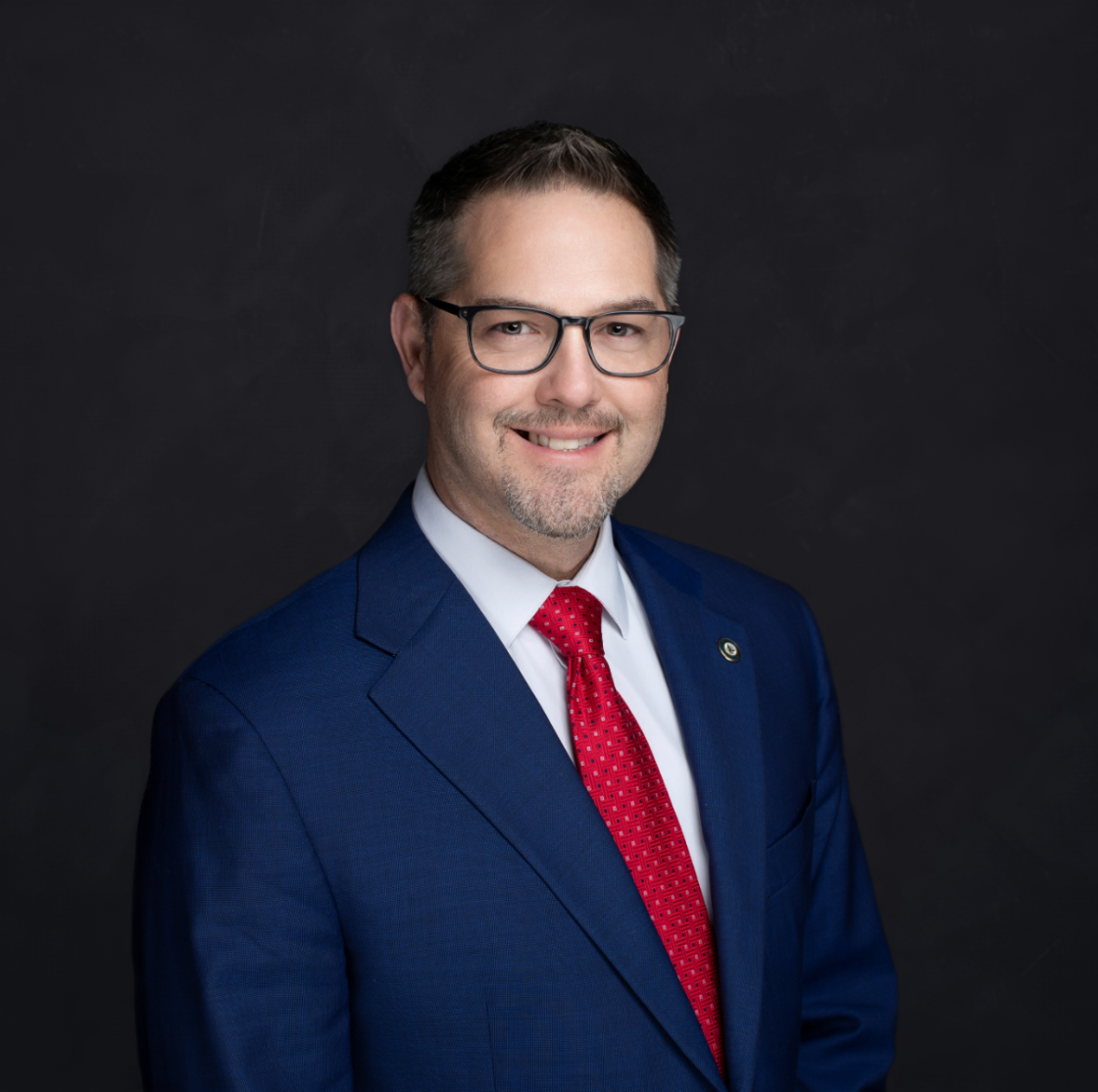 Professional headshot of a man with glasses, wearing a blue suit, white shirt, and red tie, smiling against a dark background.