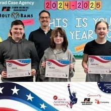 Four young women and a man holding certificates on a stage with a patriotic backdrop, celebrating a graduation or award ceremony.
