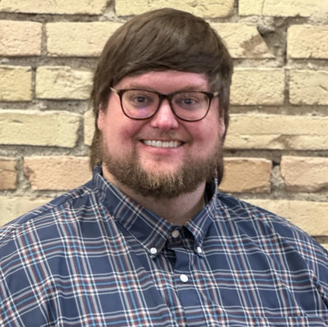 A man with glasses, a beard, and brown hair smiling, wearing a blue plaid shirt, in front of a brick wall.
