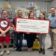 Group of five people standing in a store aisle holding a large charity check for $5,000, smiling, with shelves of products behind them.