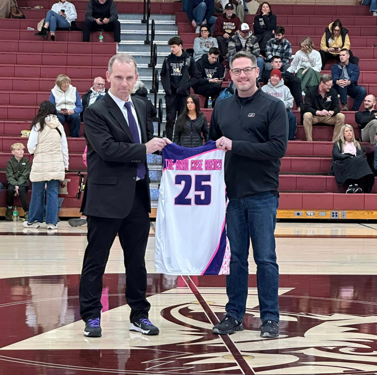 Two men are standing on a basketball court, holding a white and purple basketball jersey with the number 25 and the words "THE BRAD CASE AGENCY" printed on it. They are smiling and facing the camera. The man on the left is wearing a black suit with a white shirt and purple tie, while the man on the right is dressed in casual black clothing and glasses. The gymnasium has red bleachers with spectators seated in the background.