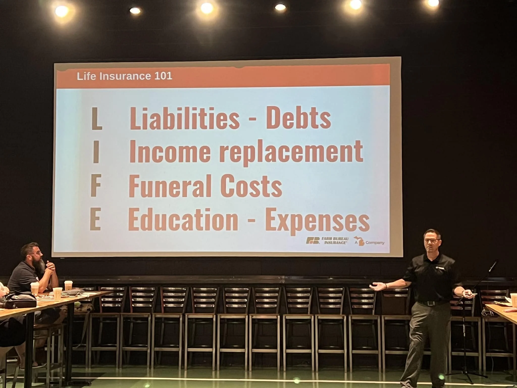 A presentation slide titled 'Life Insurance 101' displays the acronym 'LIFE' with each letter standing for Liabilities, Income replacement, Funeral Costs, and Education - Expenses. A presenter stands to the right, gesturing towards the slide, while attendees sit at tables with coffee cups and papers.