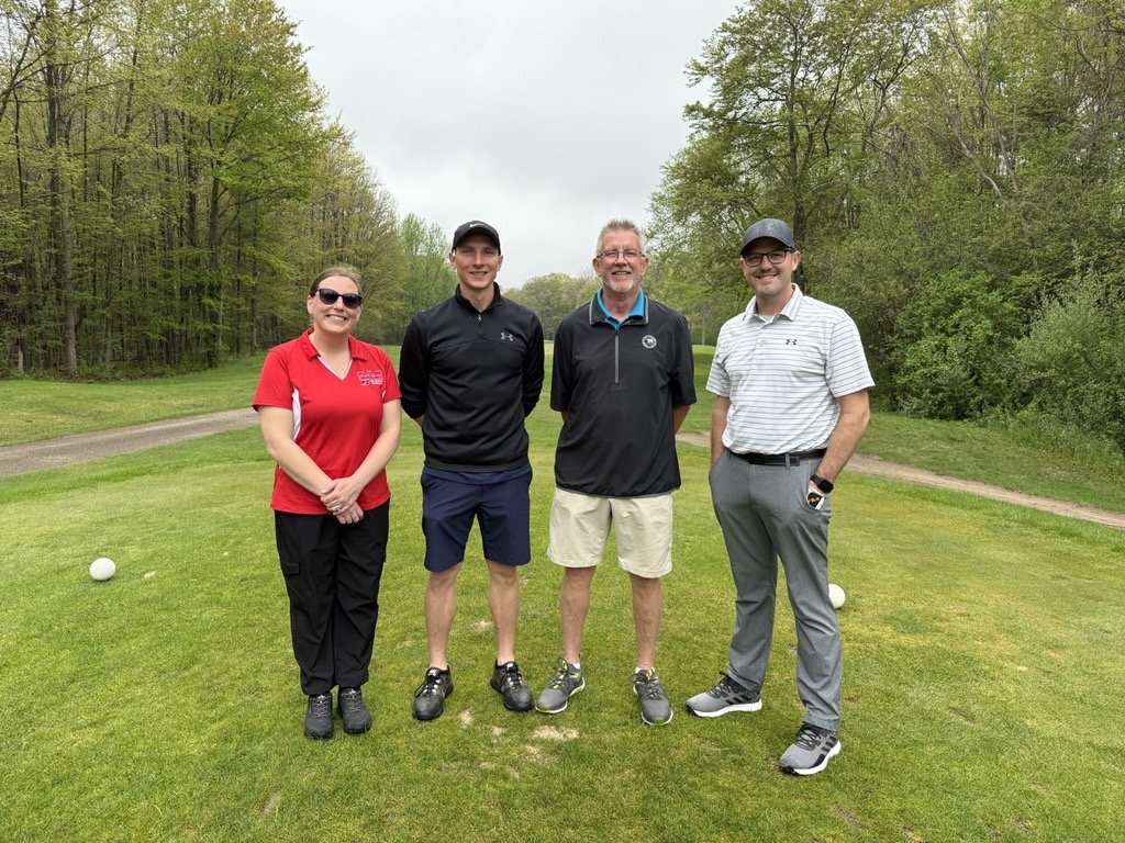 Four people standing on a golf course with trees in the background, smiling, dressed in golf attire.